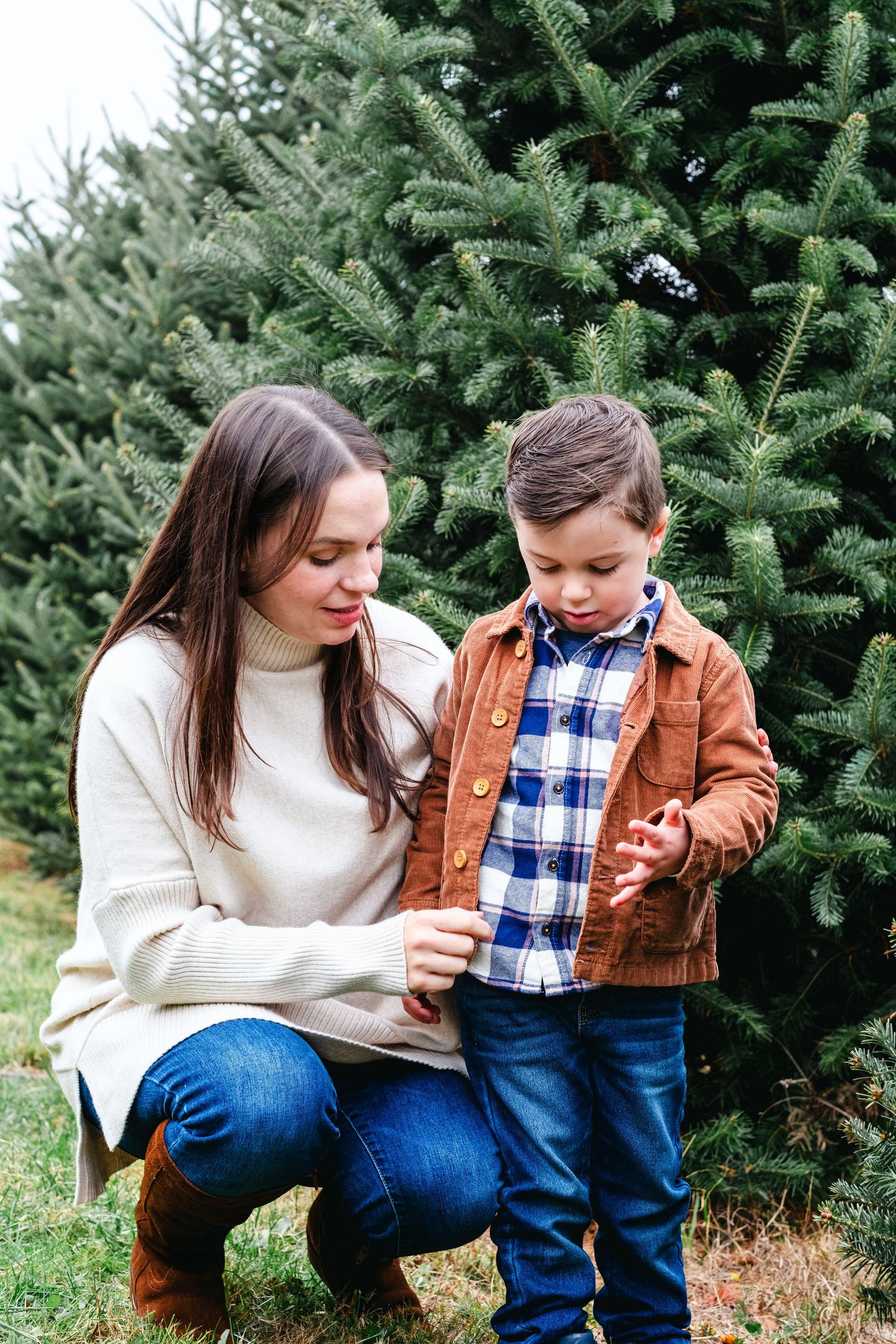 A mother crouches beside her young son at a Christmas tree farm, both dressed in casual winter attire among evergreen trees.