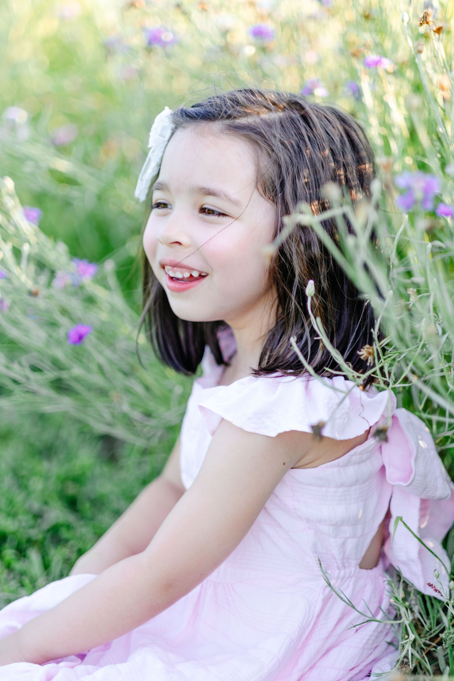 Young girl in pink dress sitting in wildflower meadow, smiling joyfully with white bow in dark hair.
