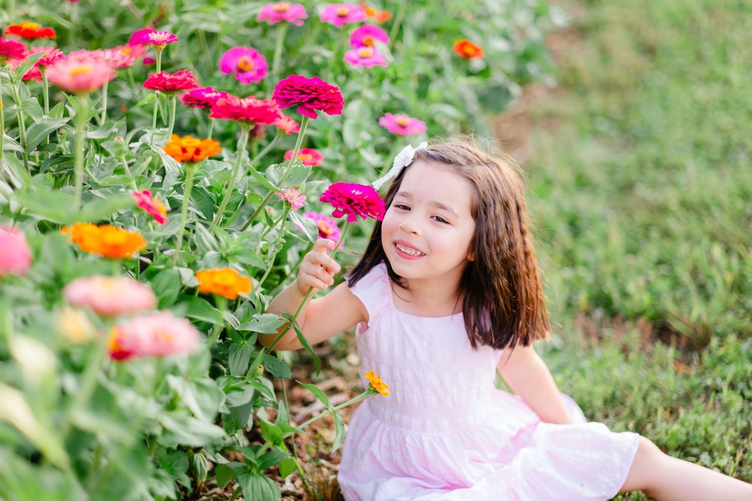 A smiling young girl in a pink dress sits among colorful zinnia flowers in a garden, reaching out to touch the blooms.