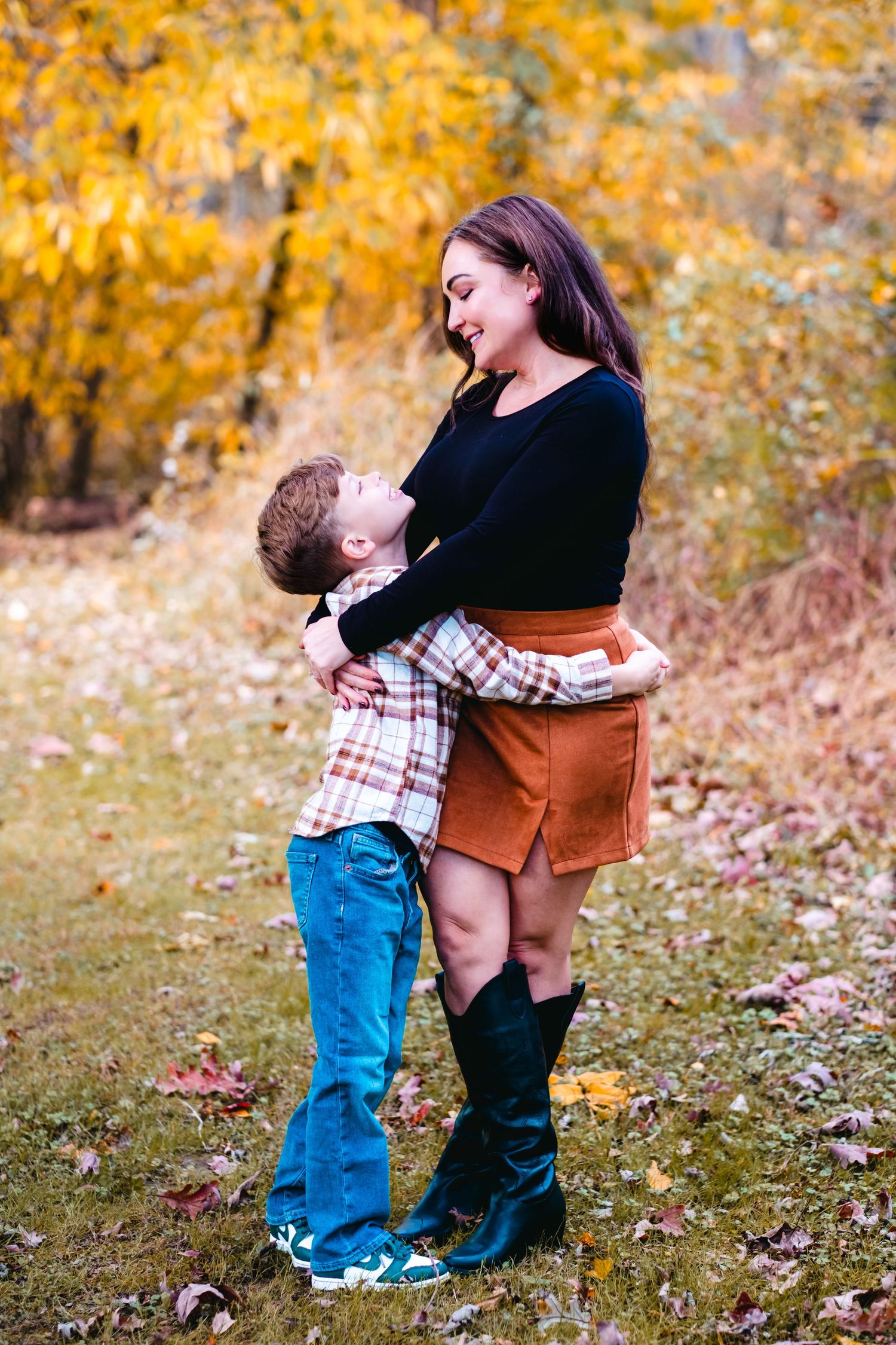 A woman and young boy share a warm embrace in an autumn forest setting with golden foliage.