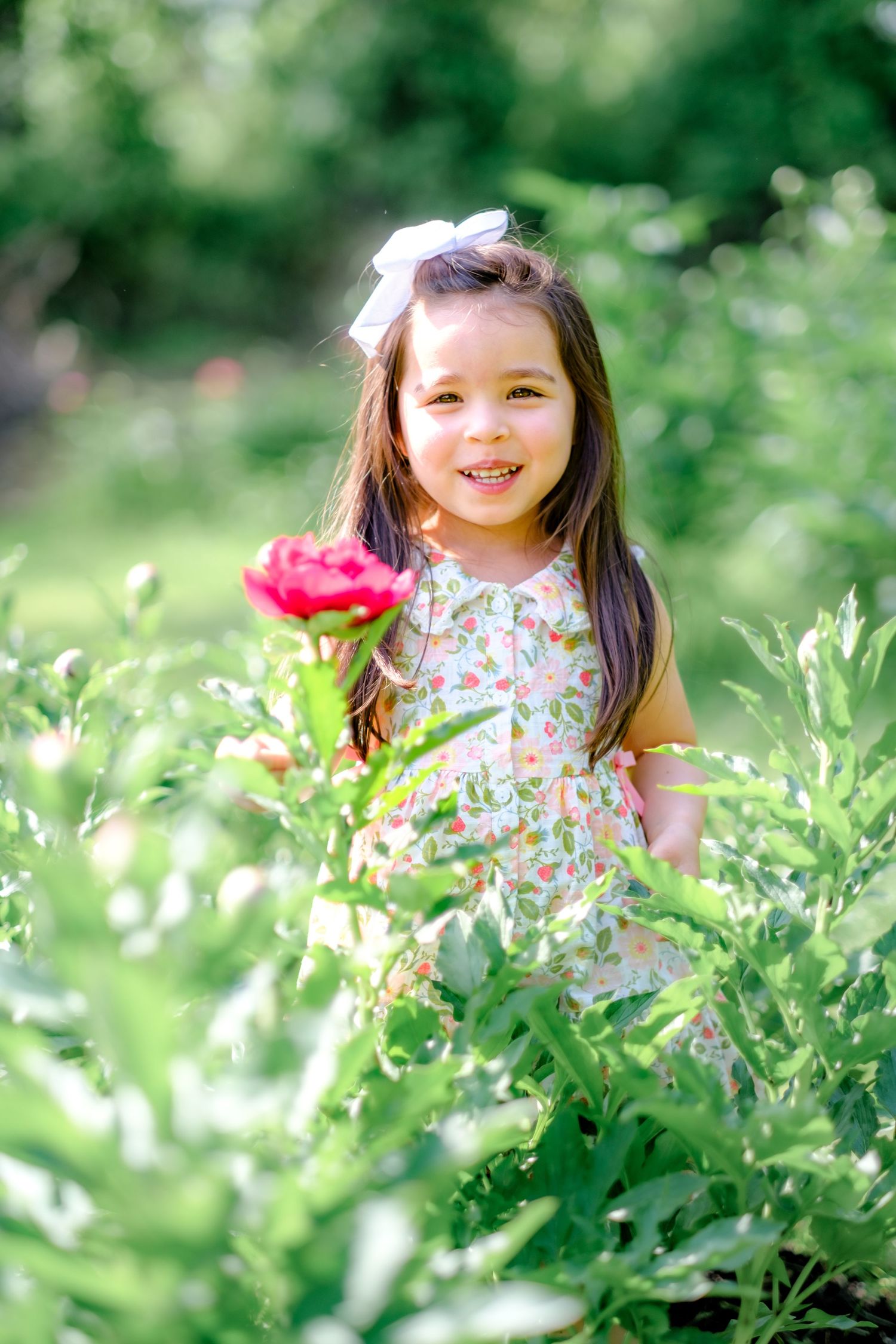 Smiling little girl in floral dress with white bow in hair standing among green plants and pink flower outdoors.