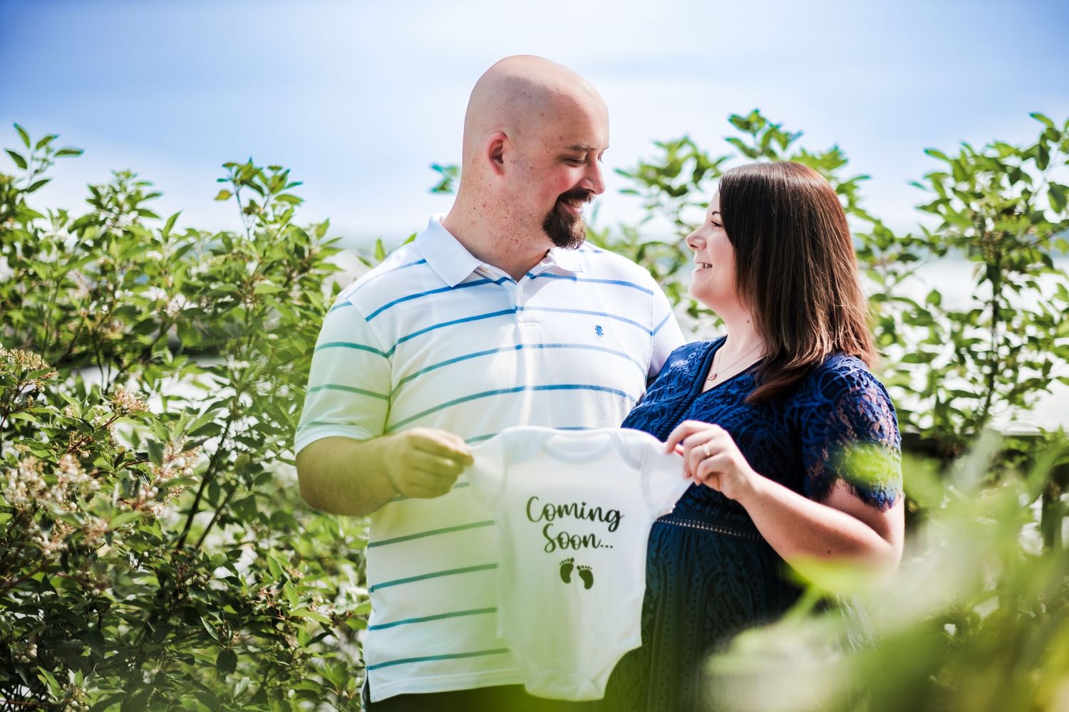Couple holding a Coming Soon baby onesie outdoors, announcing pregnancy among lush green trees on a sunny day.