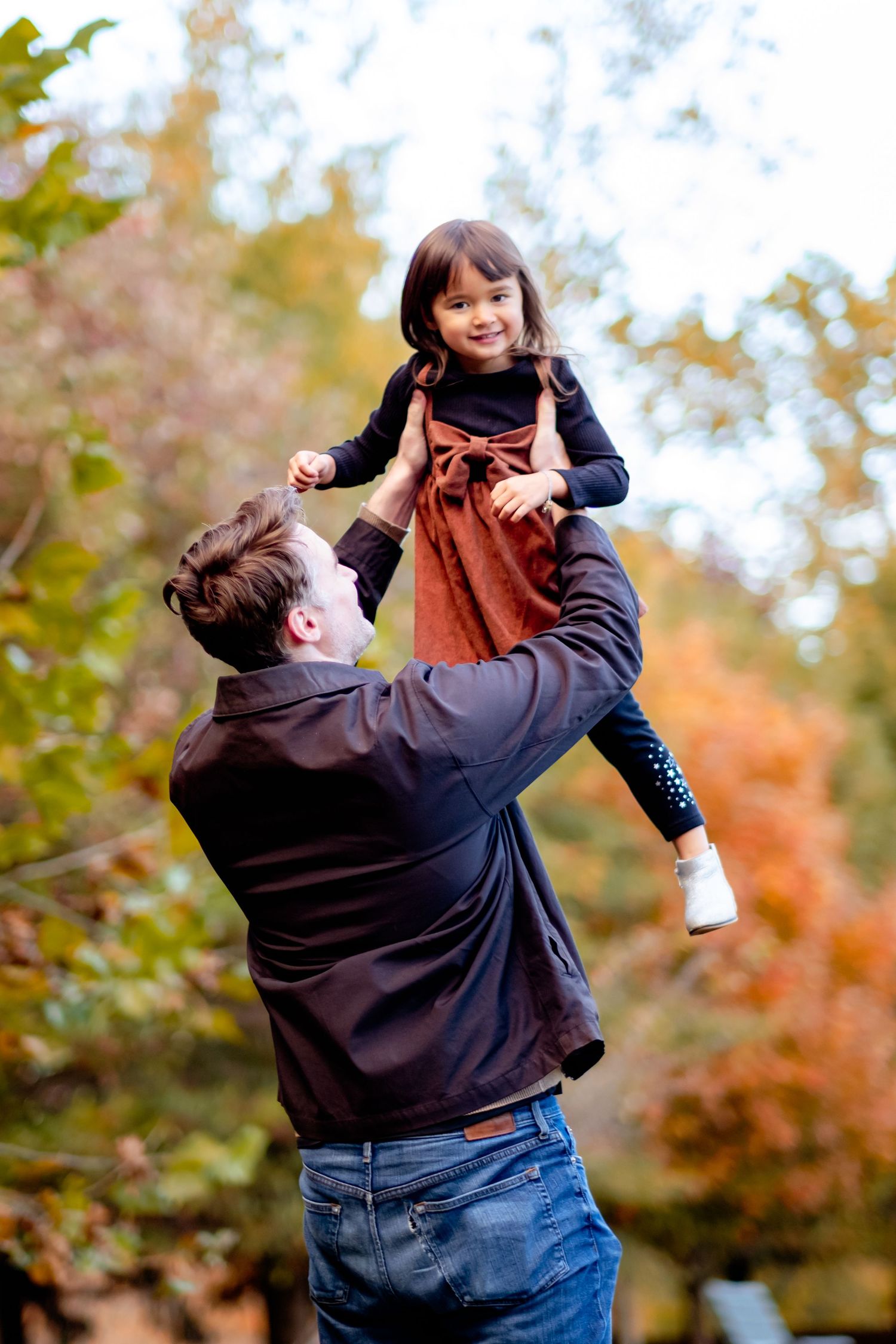 Father lifting smiling daughter in autumn park, surrounded by colorful fall foliage.