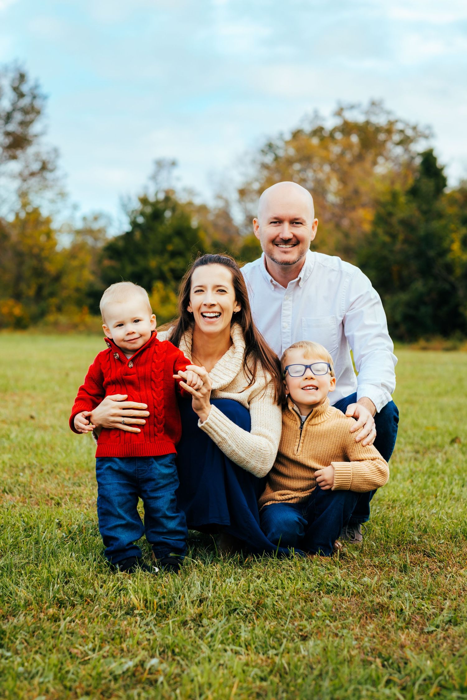 Family of four posing outdoors in fall, parents smiling with two young boys in colorful autumn sweaters on green grass.