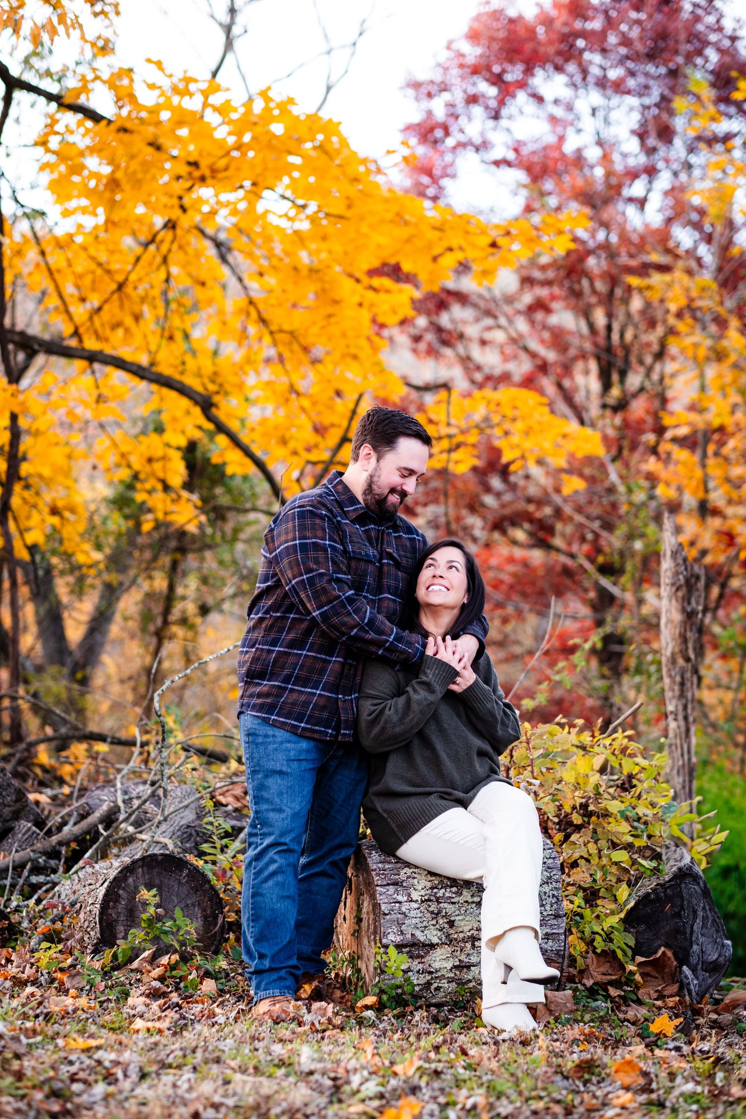 Couple embracing among vibrant autumn foliage, woman seated on log smiling up at bearded man in plaid shirt.
