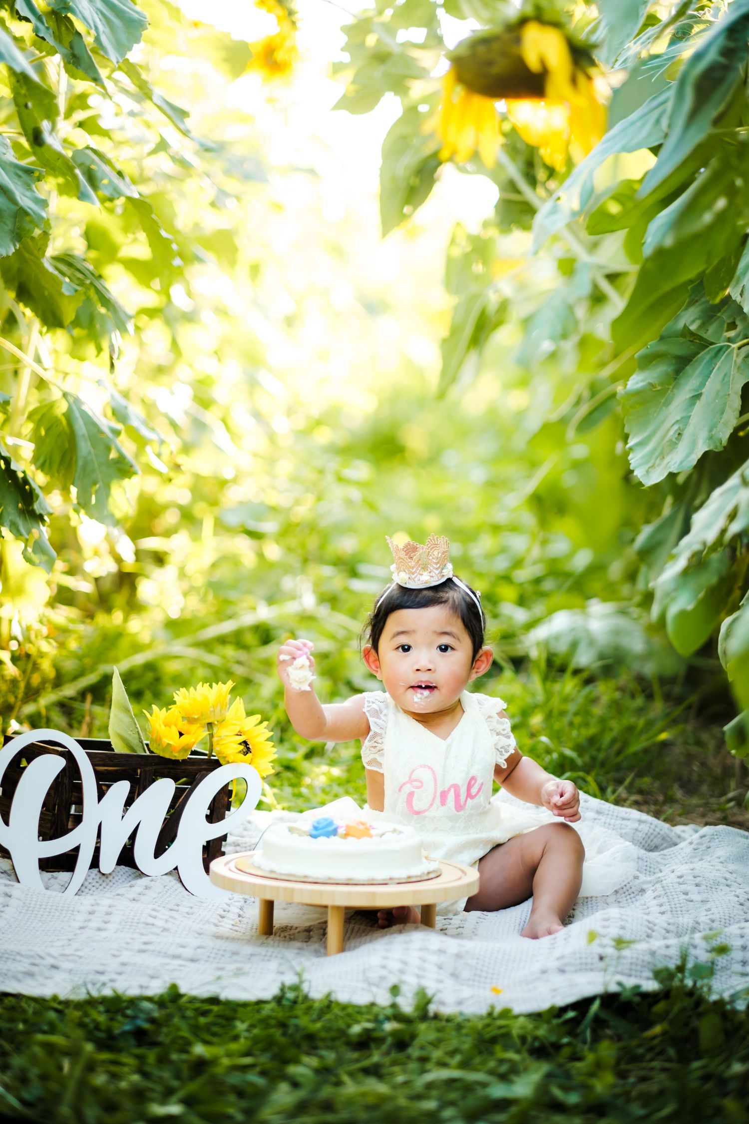 Baby girl in white dress celebrates first birthday with cake among sunflowers in a sunny garden setting.