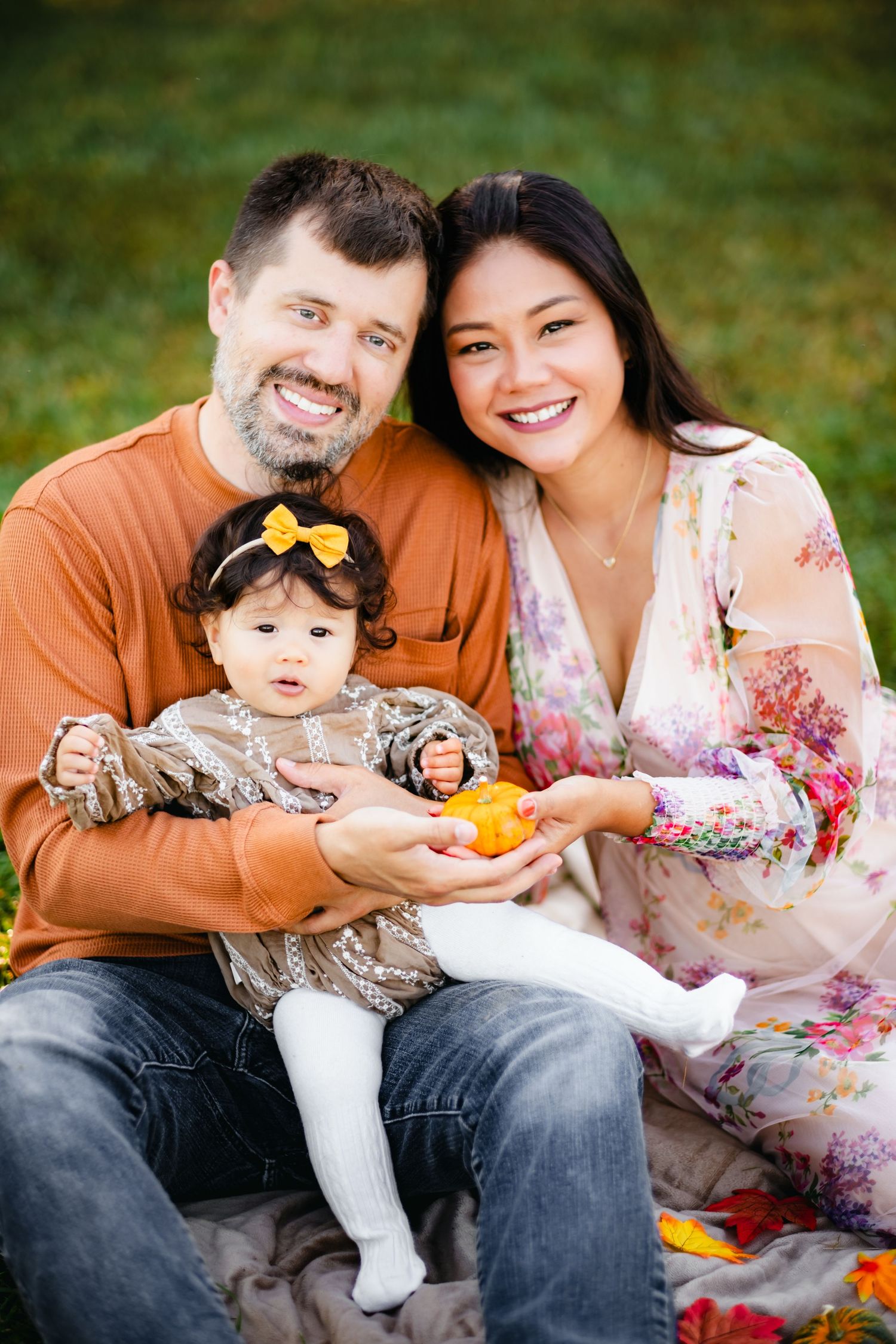 Happy interracial family sitting outdoors in autumn, parents smiling and holding baby girl with yellow bow headband.