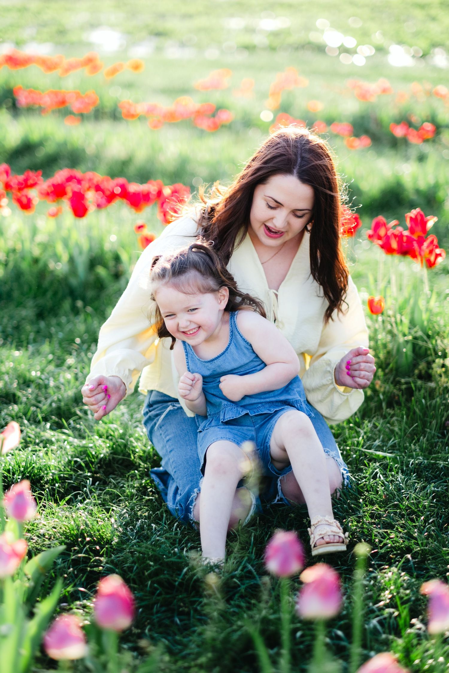 Mother and daughter laughing together in a vibrant tulip field, surrounded by red and pink flowers on a sunny day.