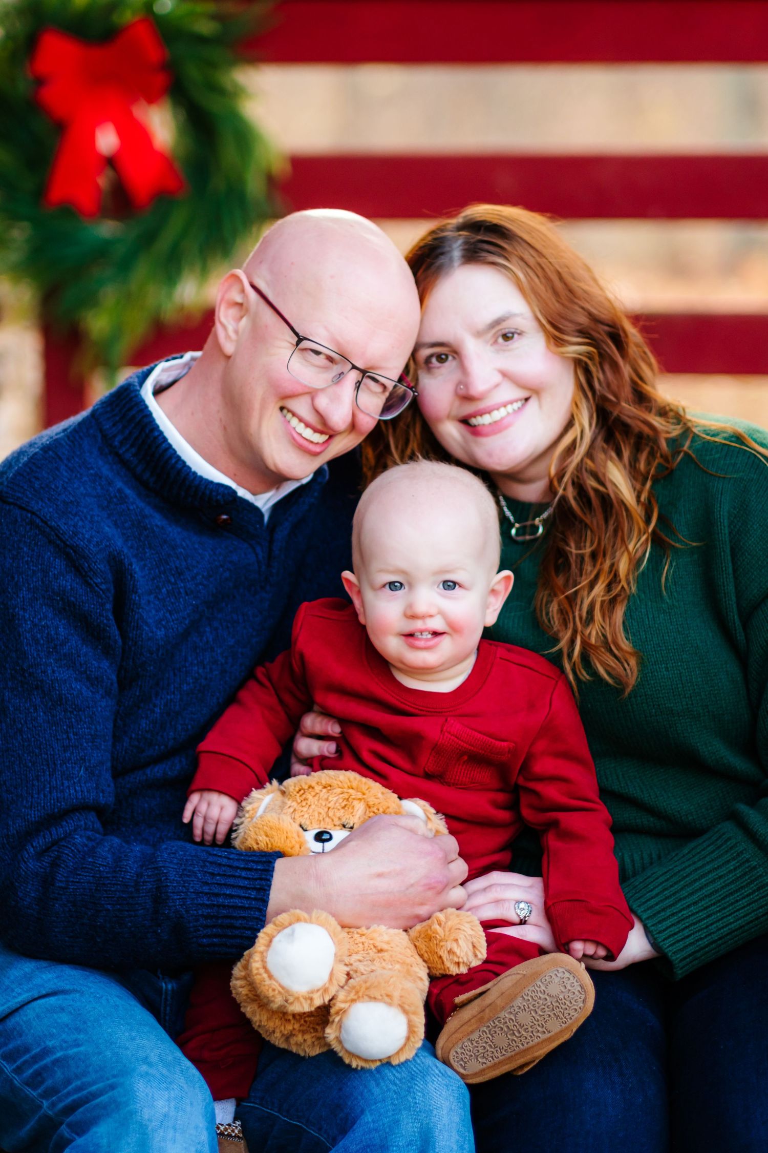 Family Christmas portrait with parents and baby holding teddy bear, festive wreath background.