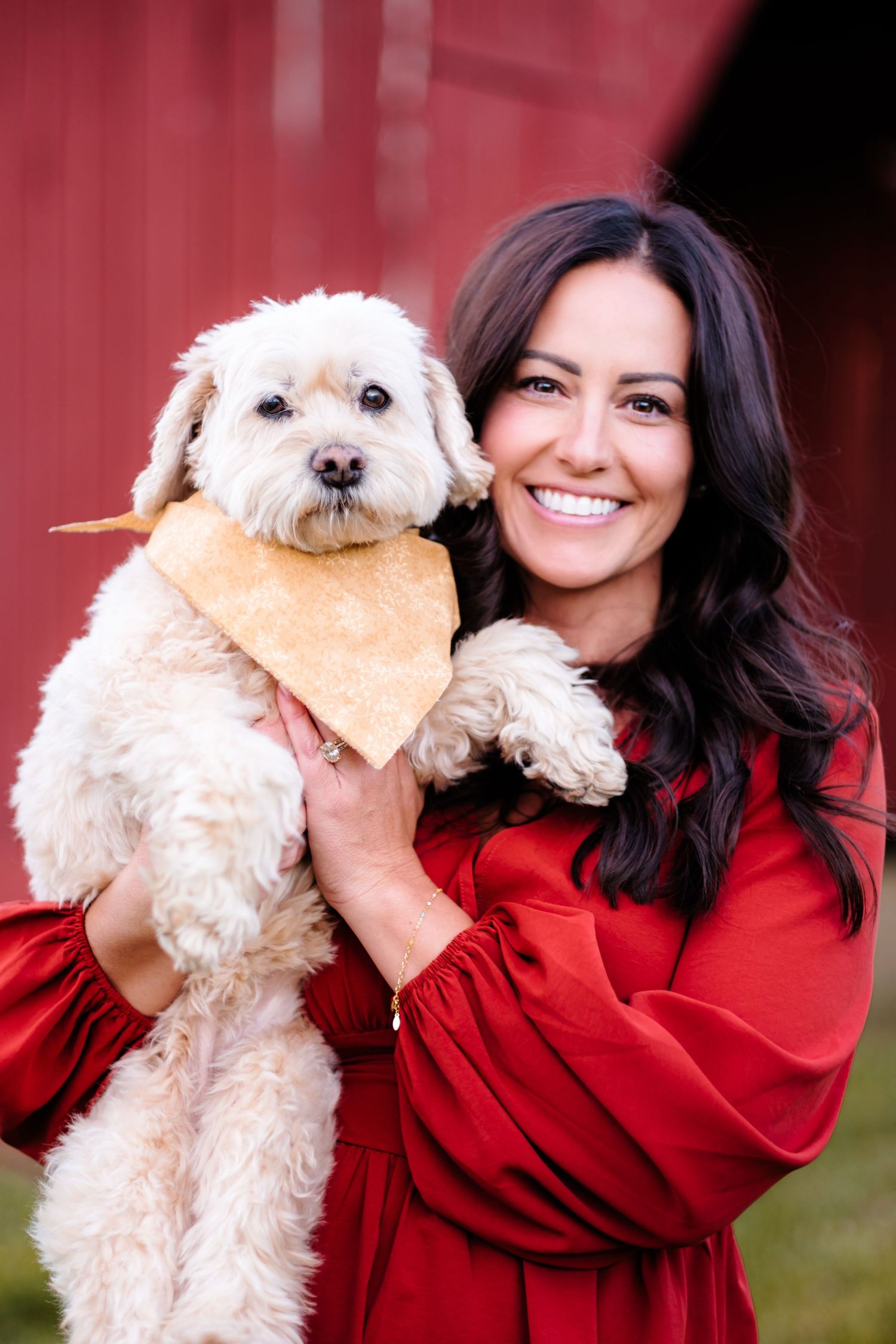 Smiling woman in red dress holding a fluffy white dog wearing a yellow bandana in front of a red barn.