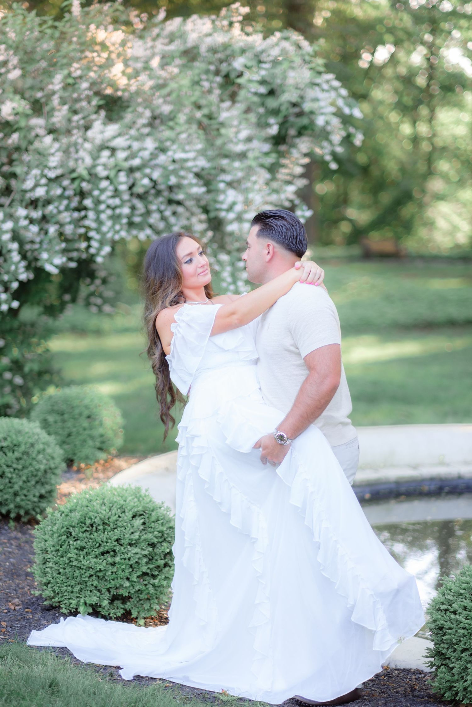 Couple in white attire embracing in a lush garden with blooming trees and manicured hedges near a pond.