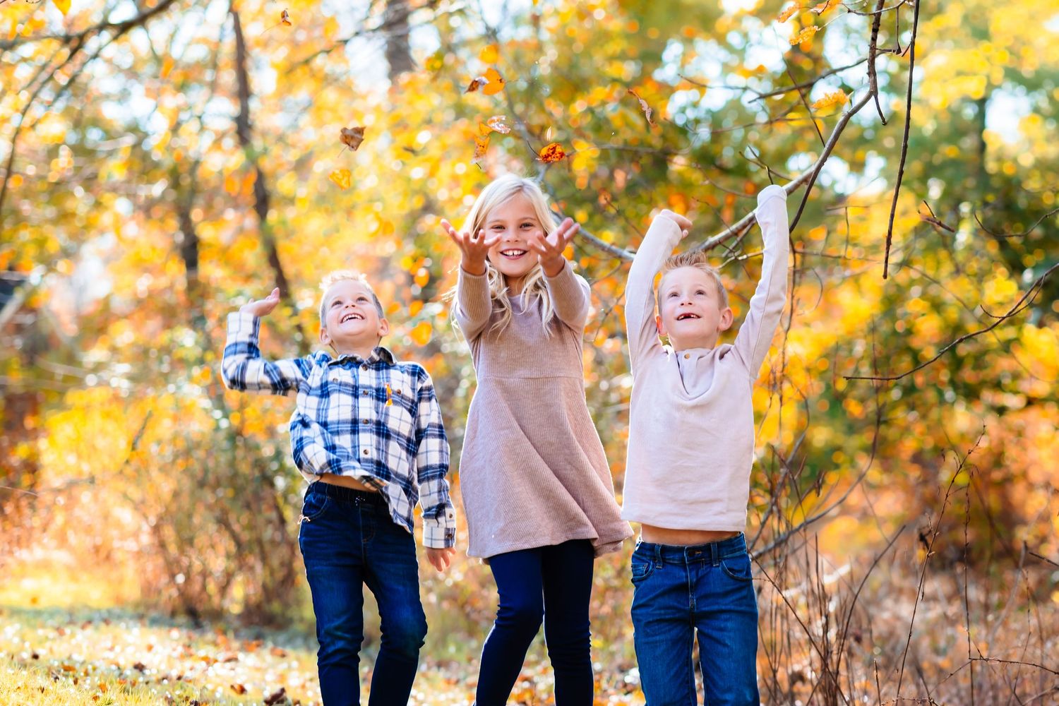 Three happy children playing with autumn leaves in a colorful fall forest, smiling and reaching up joyfully.