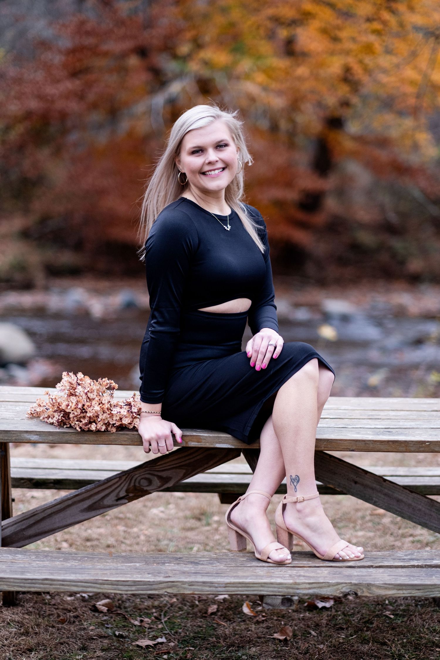 Smiling blonde woman in black cutout dress and heels sitting on wooden picnic table with autumn foliage backdrop.