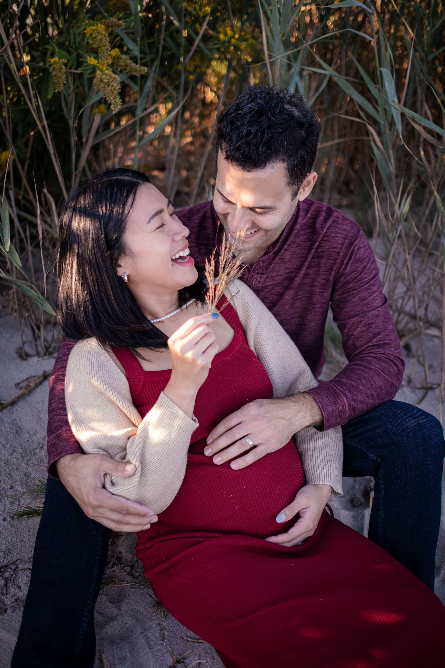 Happy pregnant couple sitting outdoors on sand, man embracing woman in red dress, sharing a joyful moment.