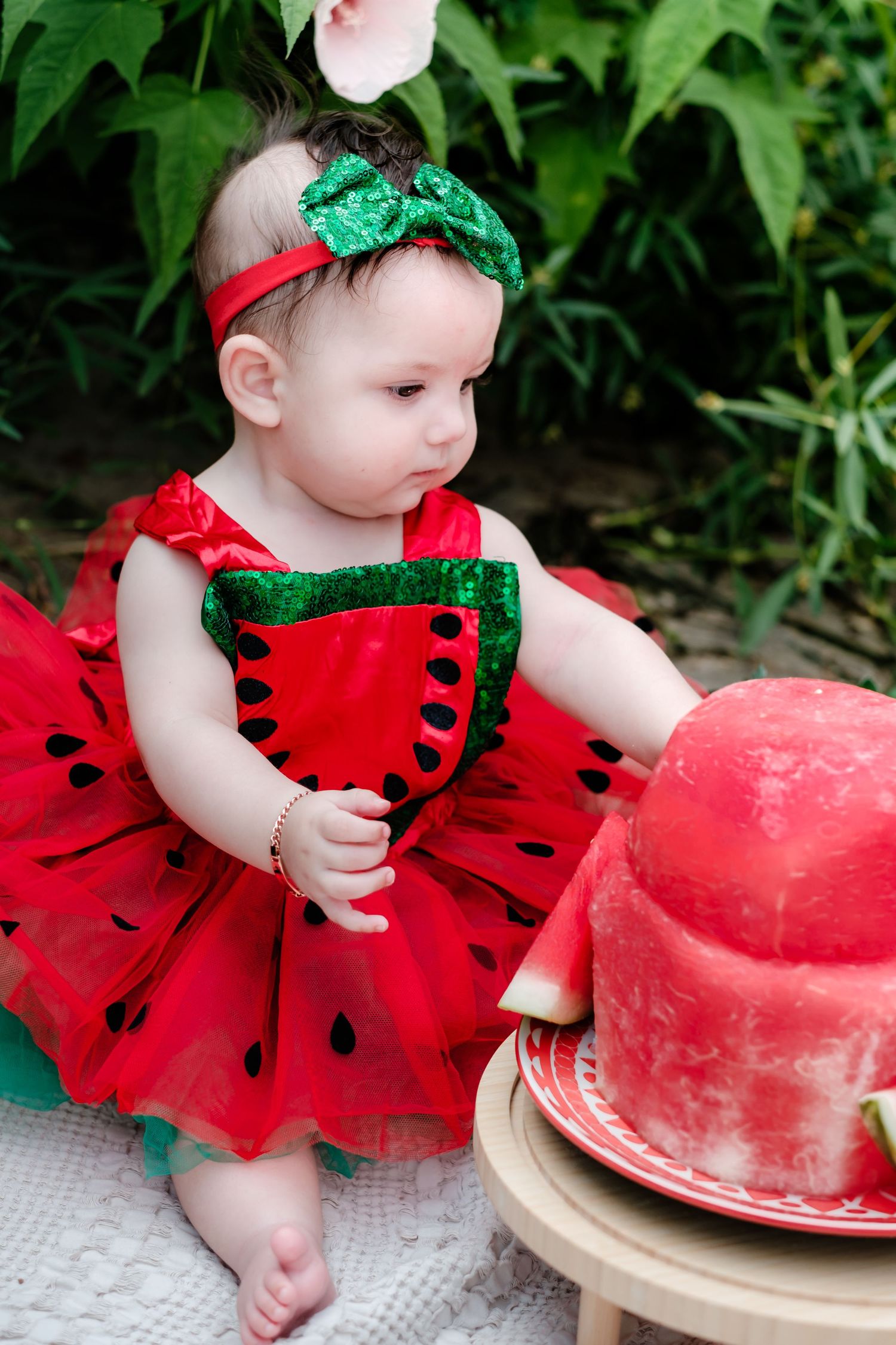 Baby girl in watermelon dress and green bow headband sits near a whole watermelon outdoors.