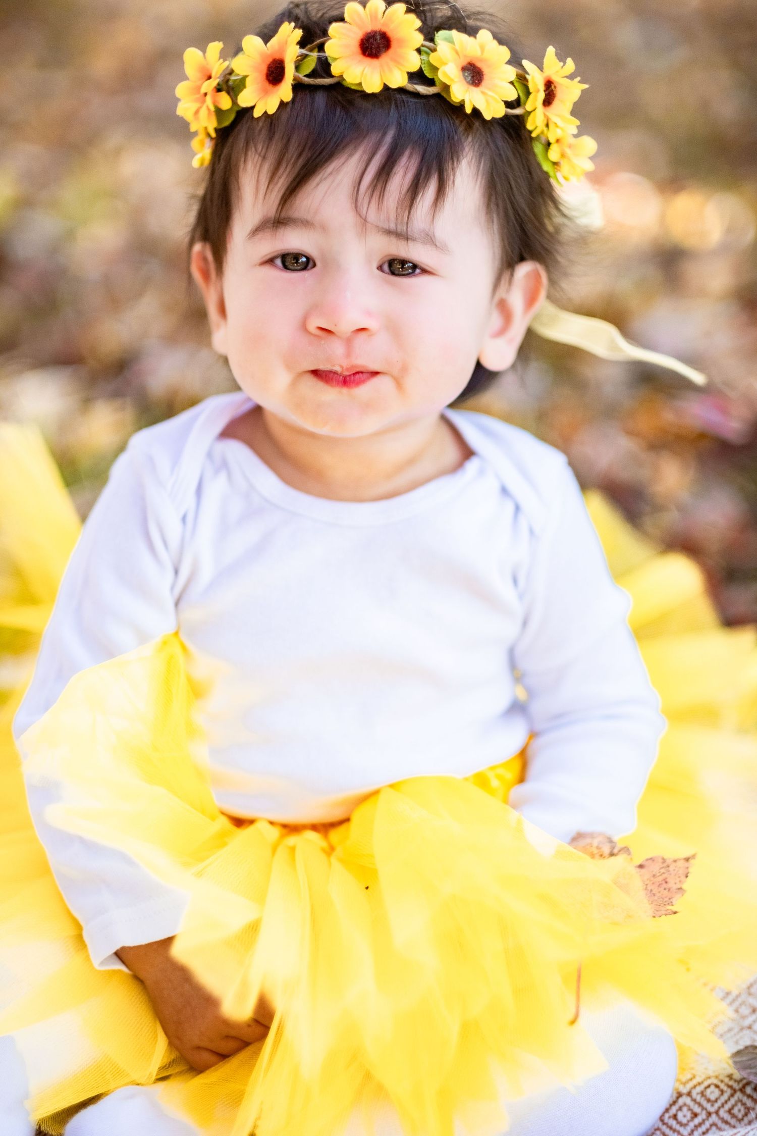 Baby girl in yellow tutu and sunflower crown sits outdoors among autumn leaves, smiling cutely.