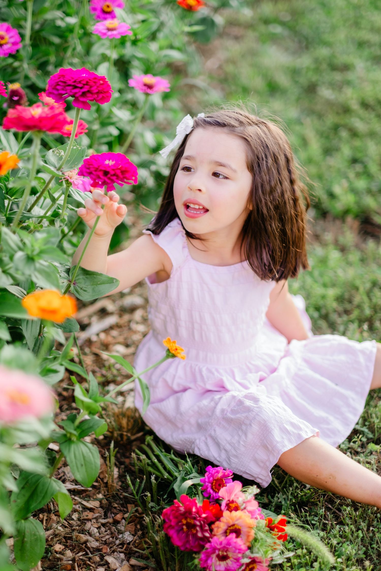 Young girl in pink dress sitting among colorful zinnia flowers in a garden, reaching out to touch a bright pink bloom.