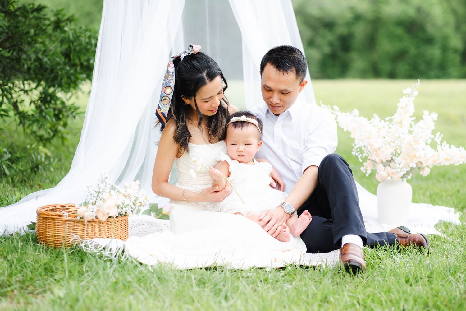 A happy Asian family with a baby girl enjoys a picnic on green grass under a white canopy with flowers.