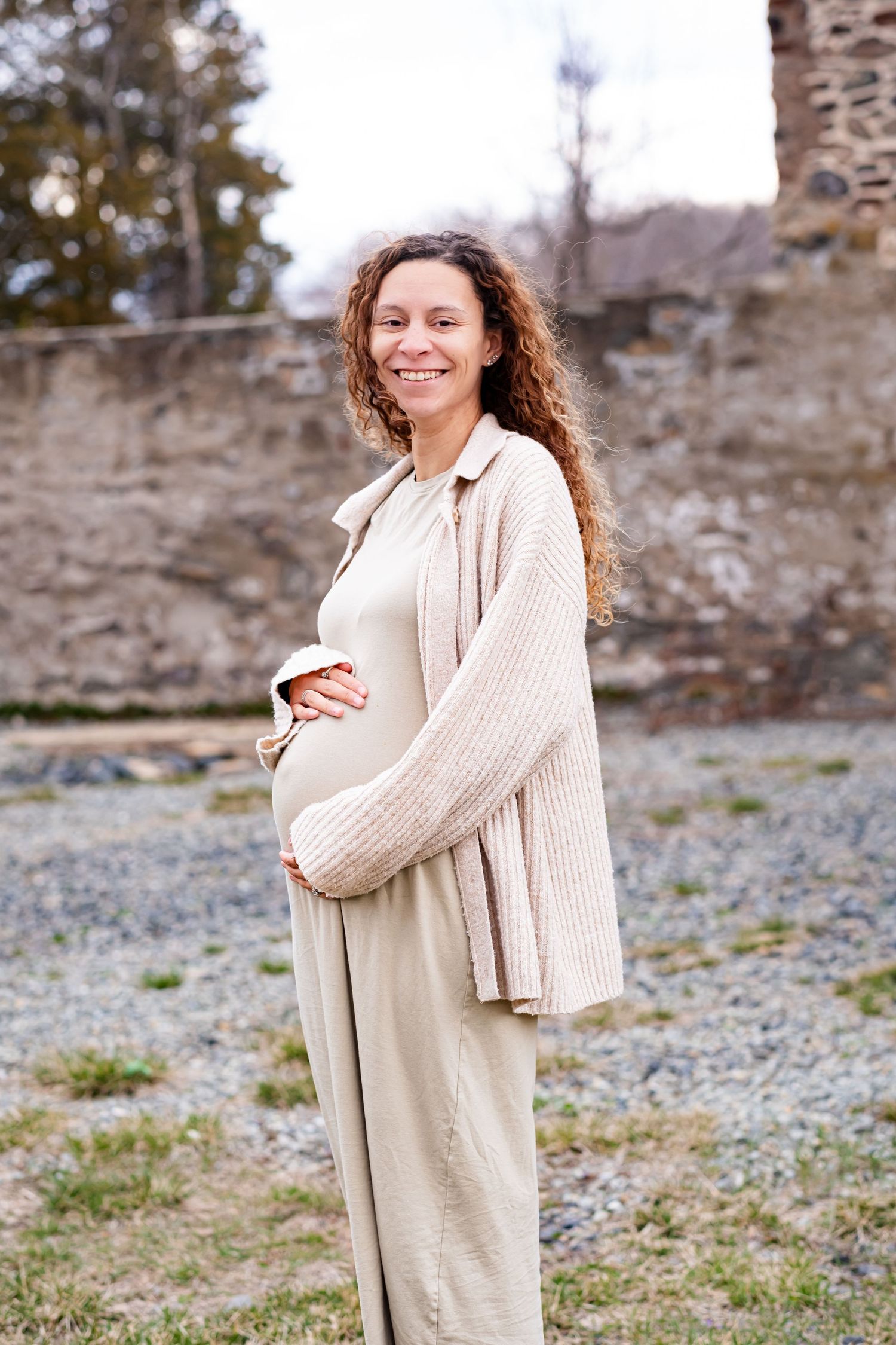 Smiling pregnant woman in beige dress and knit cardigan cradling her baby bump outdoors near a stone wall.