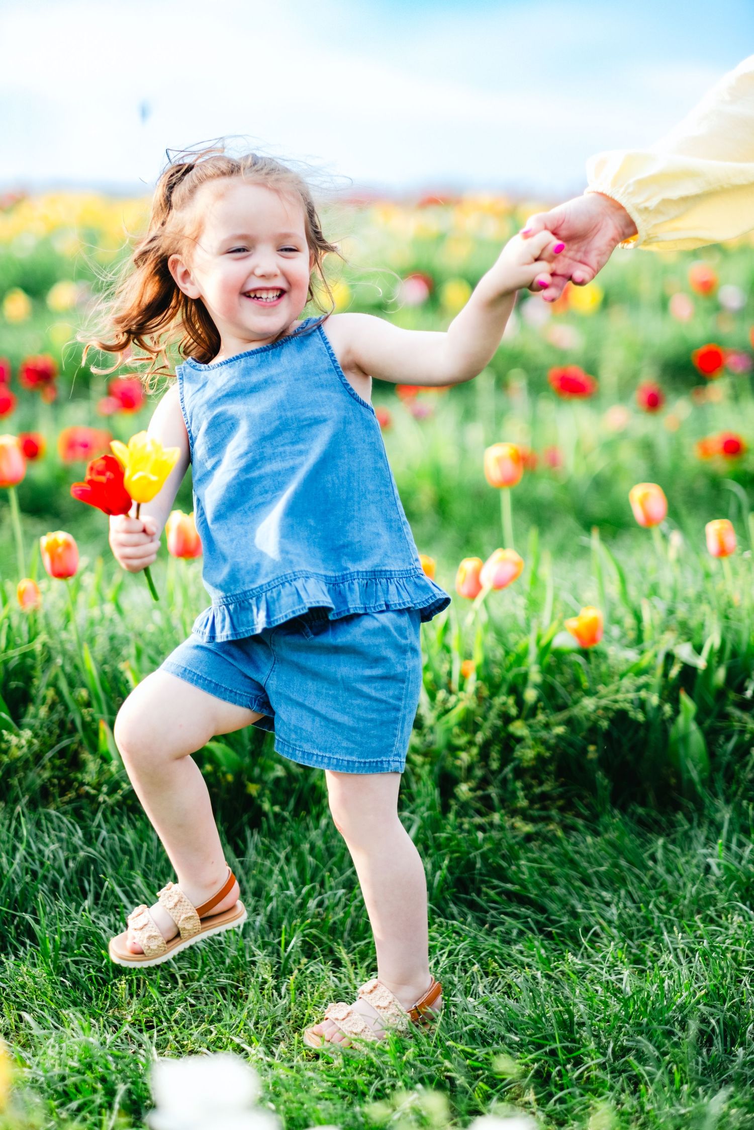 Happy little girl in blue denim dress holding colorful tulips while dancing in a vibrant spring flower field.