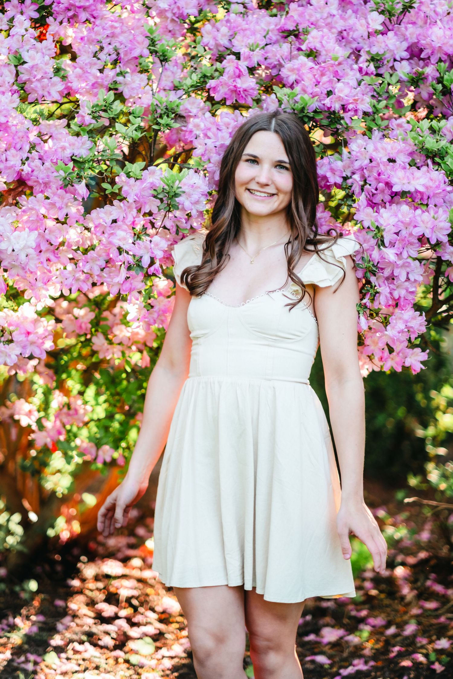 Young woman in white dress smiling among pink flowering azalea bushes in a sunny garden.