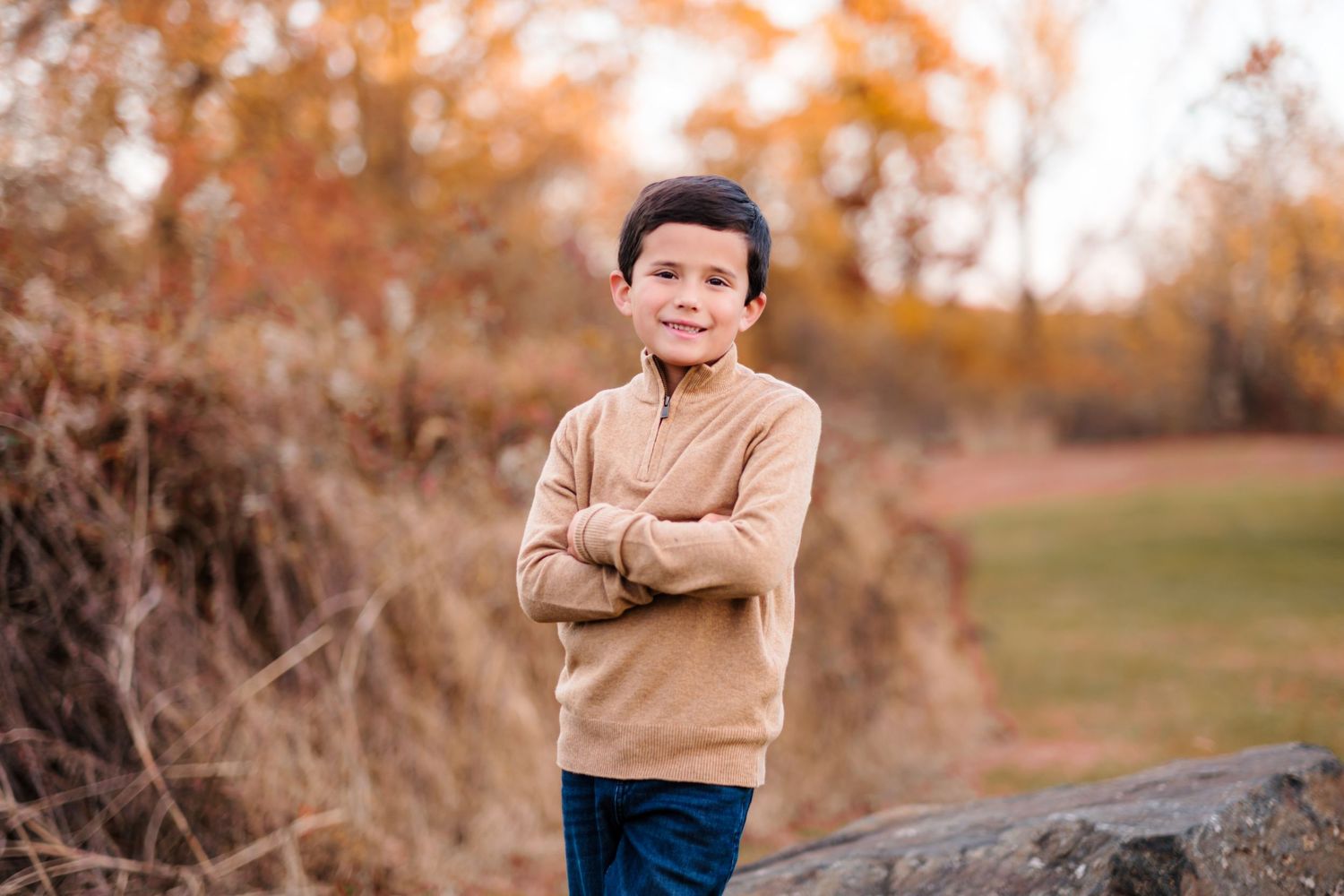A smiling boy in a tan sweater stands with arms crossed in an autumn park with colorful foliage.