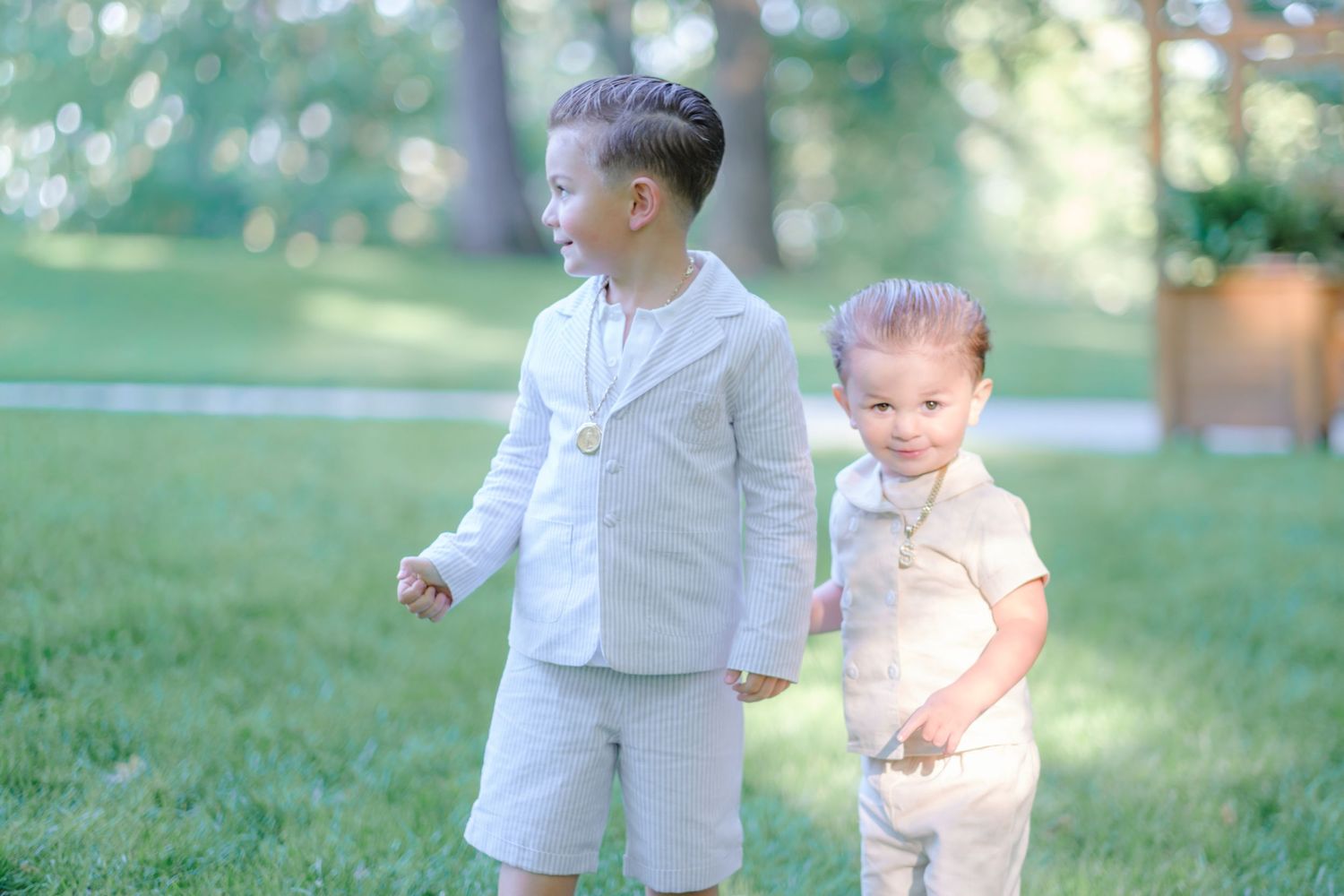 Two young boys in matching beige suits walk on green grass, smiling outdoors in a sunny park setting.