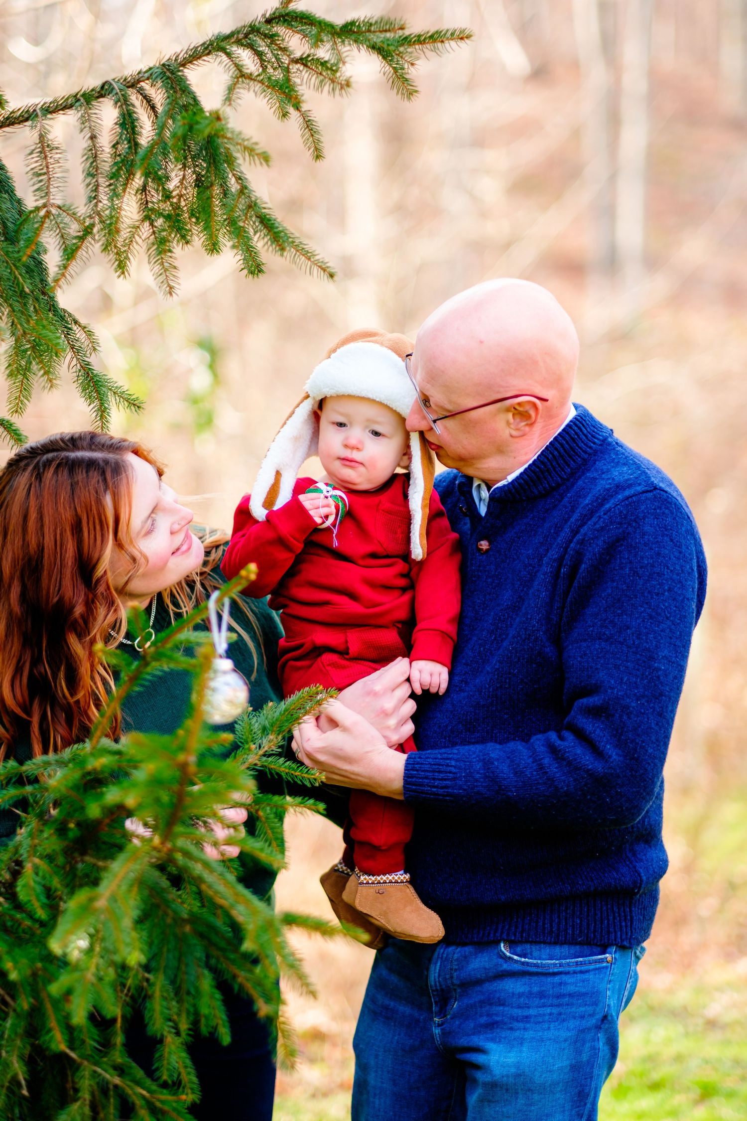 Family with baby in red outfit and bunny hat poses near decorated Christmas tree outdoors in winter setting.