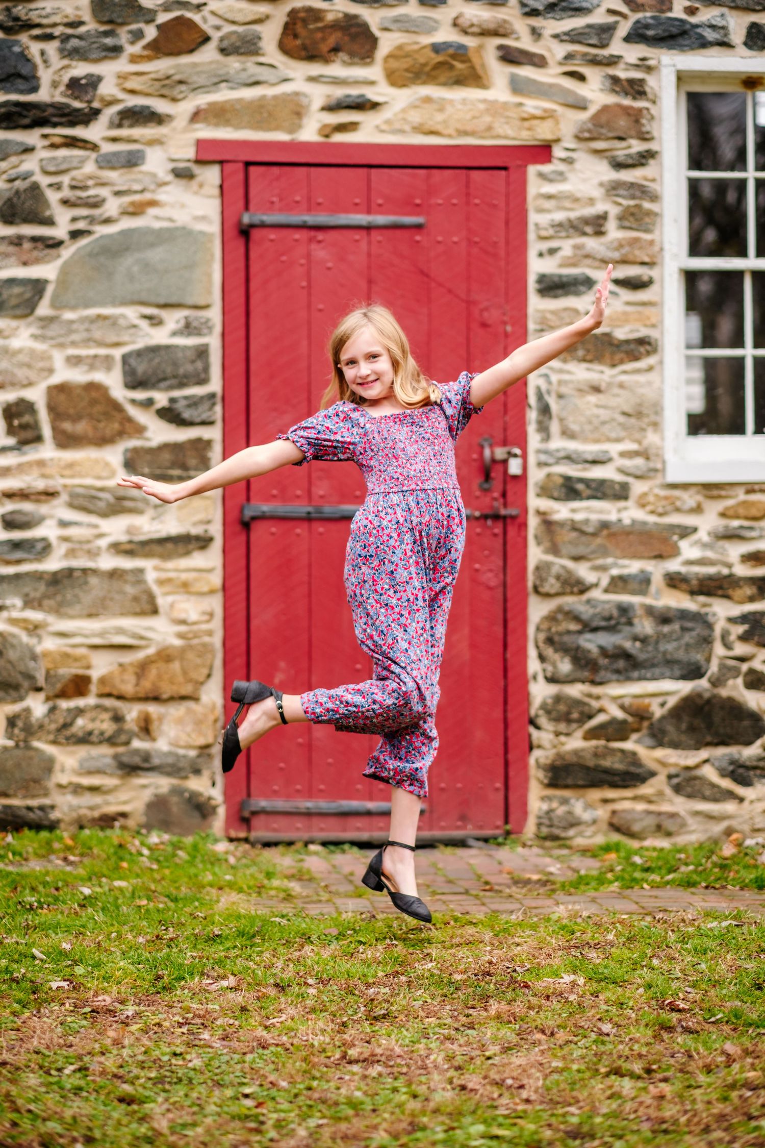 A joyful young girl in a floral jumpsuit leaps with arms outstretched in front of a rustic stone building with a red door.