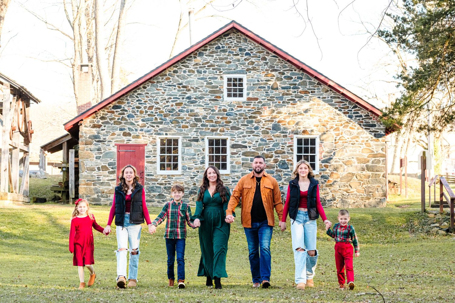 A large family of seven walks hand-in-hand across a green lawn in front of a rustic stone building.