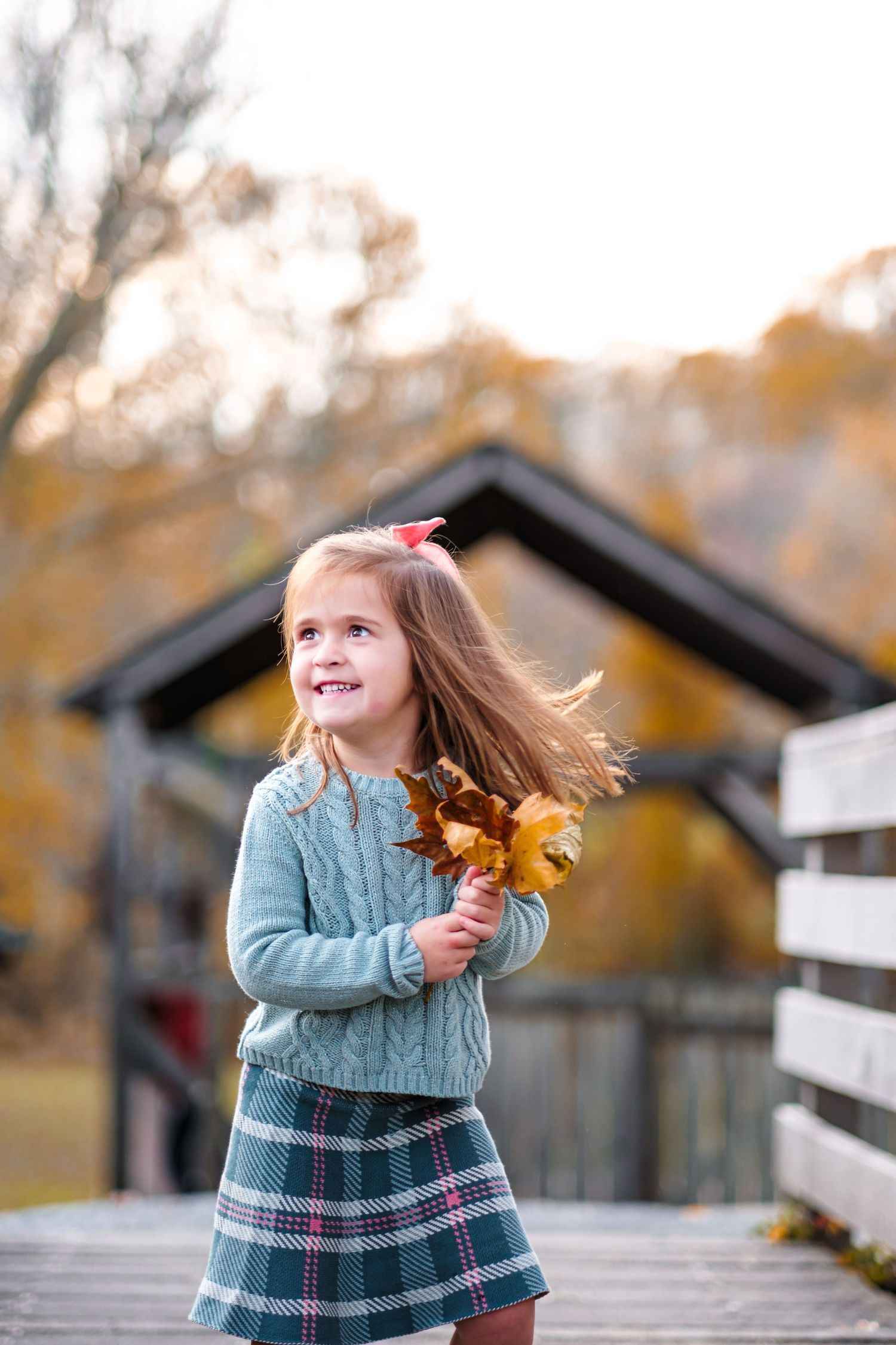 Young girl in teal sweater and plaid skirt holds autumn leaves on wooden bridge surrounded by fall foliage.