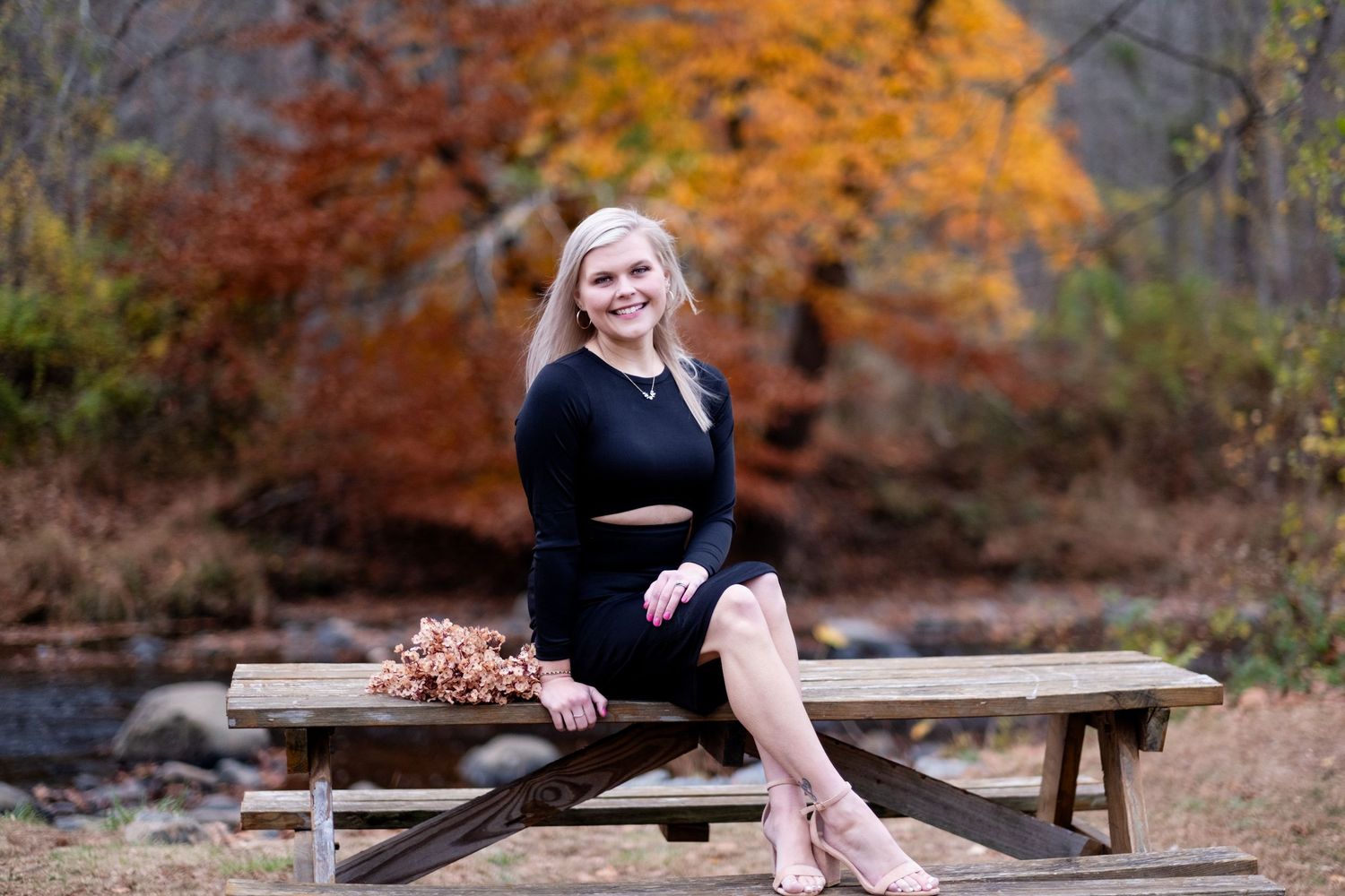 Blonde woman in black dress sitting on picnic table surrounded by colorful autumn foliage in a park setting.