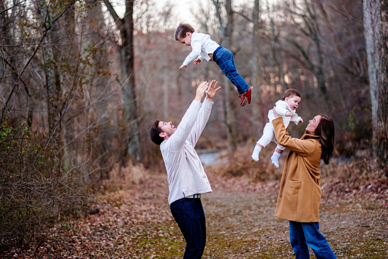 Family playing outdoors in autumn forest, parents joyfully lifting two young children in the air.