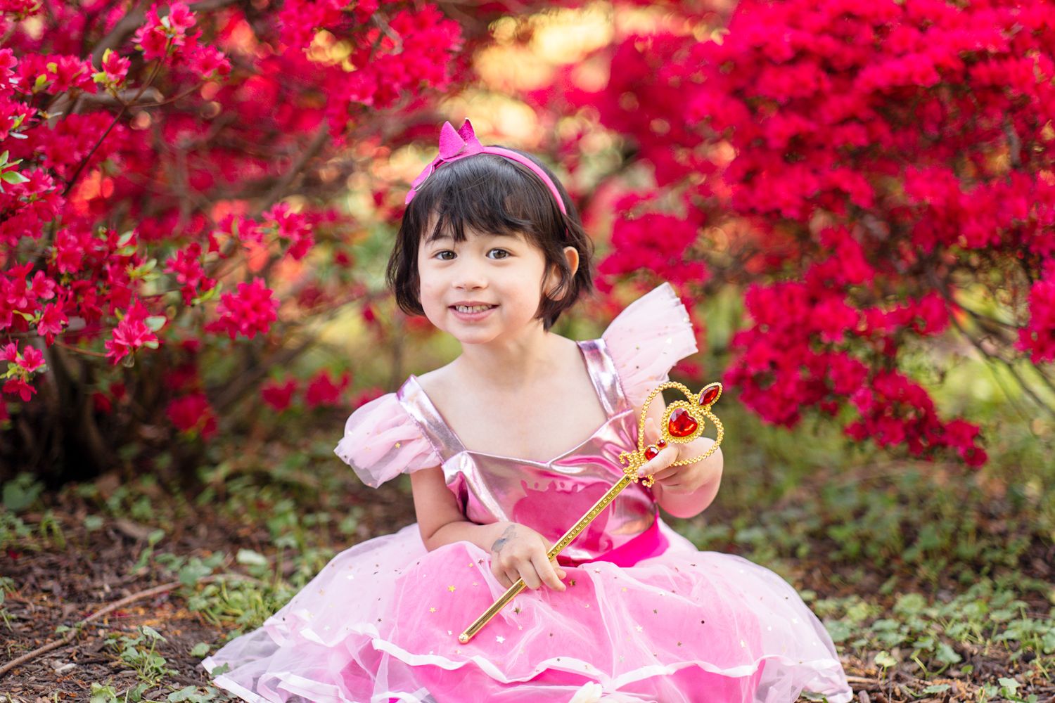 Young girl in pink princess dress holding a magic wand, surrounded by vibrant red azalea bushes outdoors.