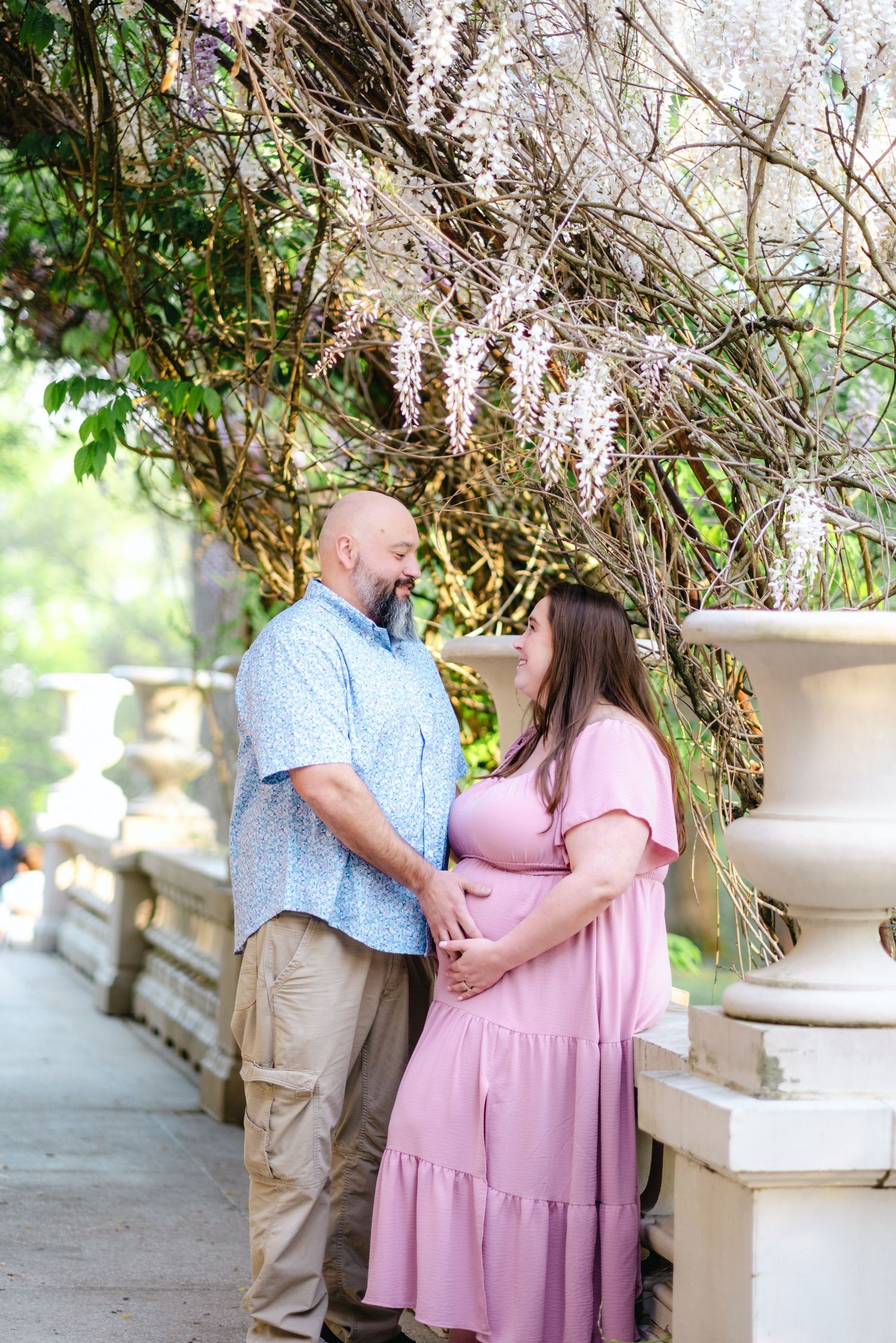 Pregnant couple in pink dress and blue shirt pose tenderly under wisteria blooms on a sunlit garden terrace.