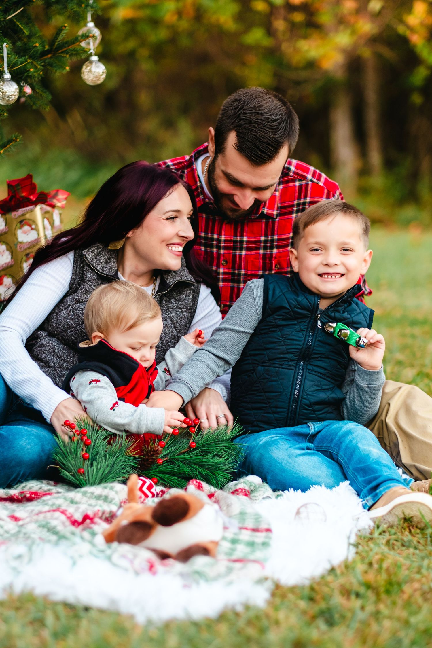 Family of four sitting on blanket under Christmas tree outdoors, smiling and enjoying holiday decorations together.