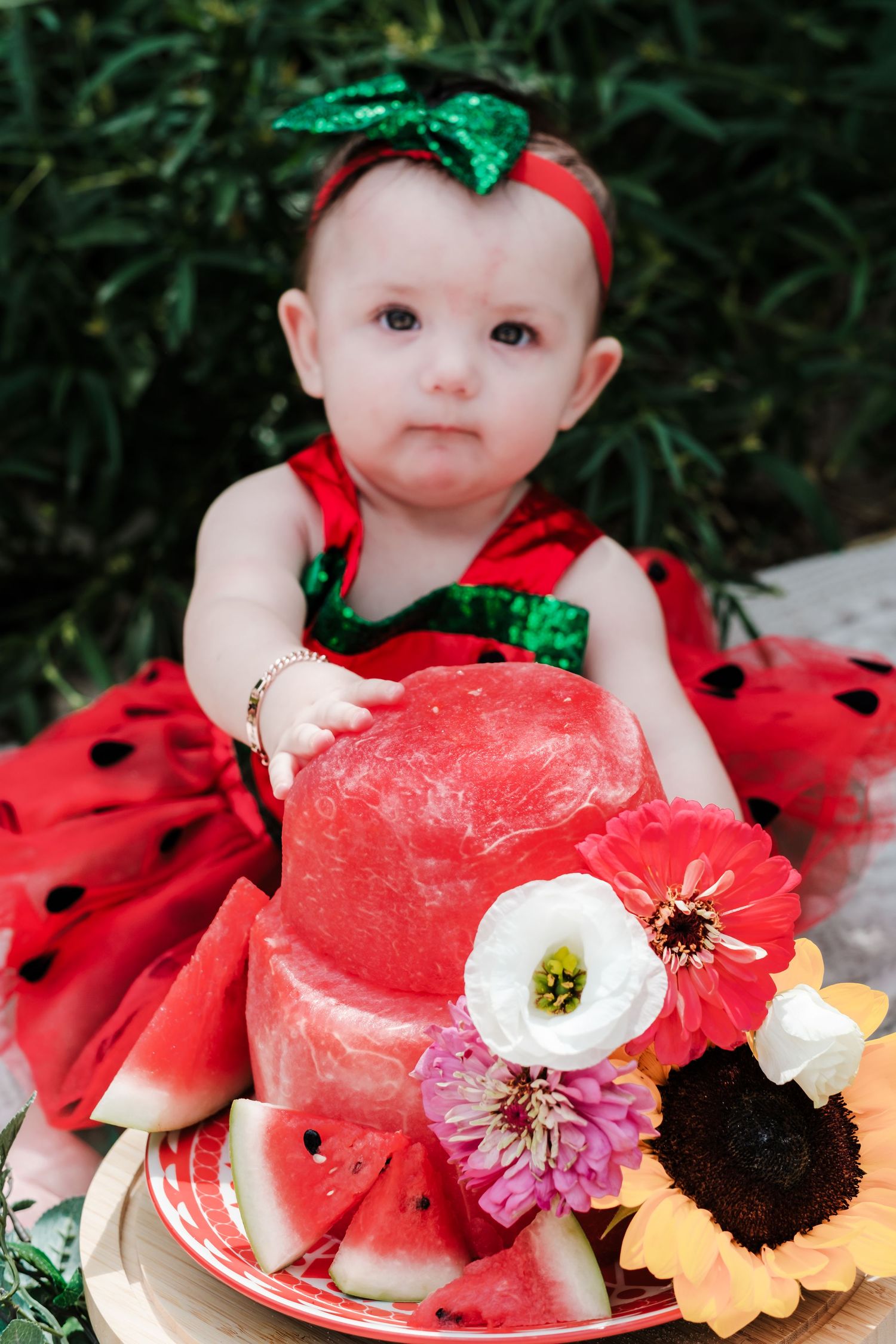 Baby girl in red ladybug dress sits with watermelon cake decorated with fresh flowers for her first birthday.