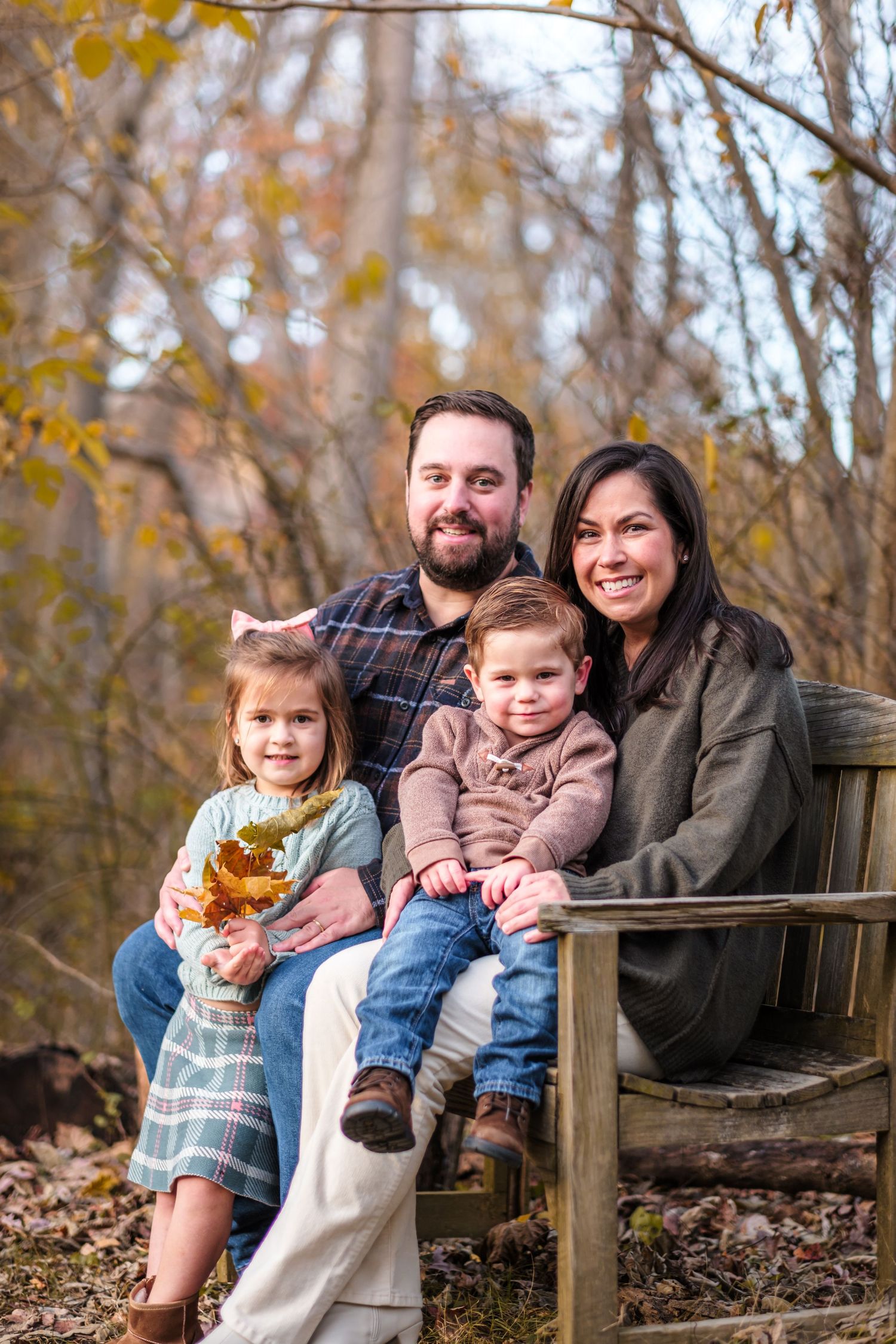 Family of four smiling on a wooden bench surrounded by autumn foliage during a fall outdoor portrait session.