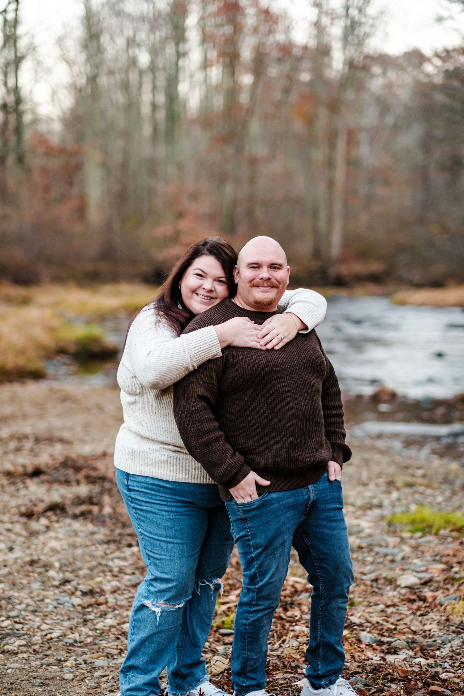 A smiling couple embraces outdoors near a stream surrounded by autumn trees, wearing casual sweaters and jeans.