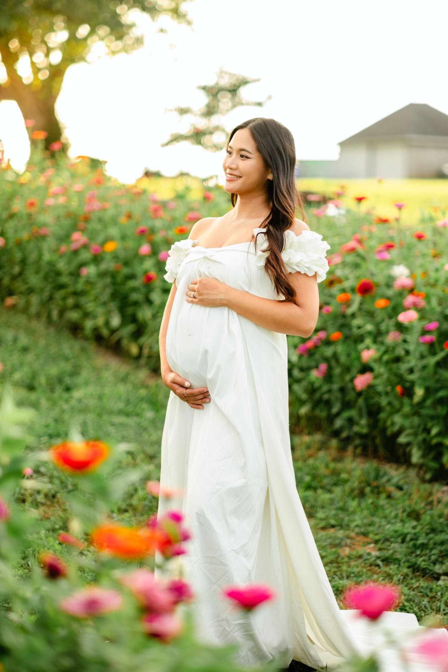 Pregnant woman in white off-shoulder gown cradling baby bump in colorful flower field at golden hour.