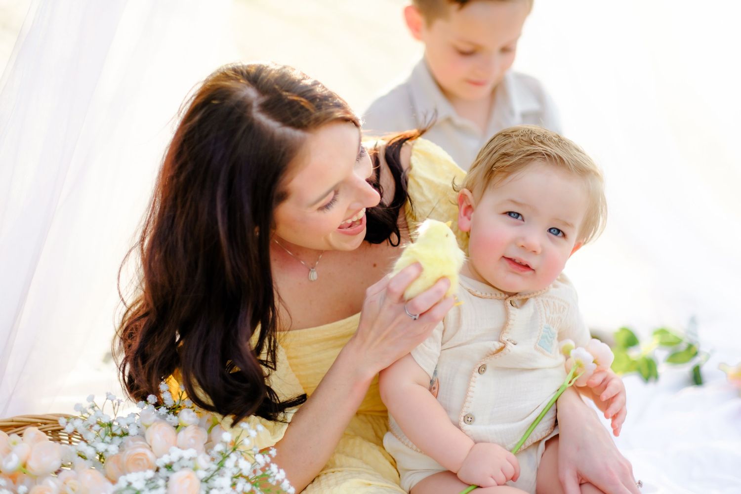 A mother in yellow dress smiles at her baby boy while an older child stands behind them indoors.