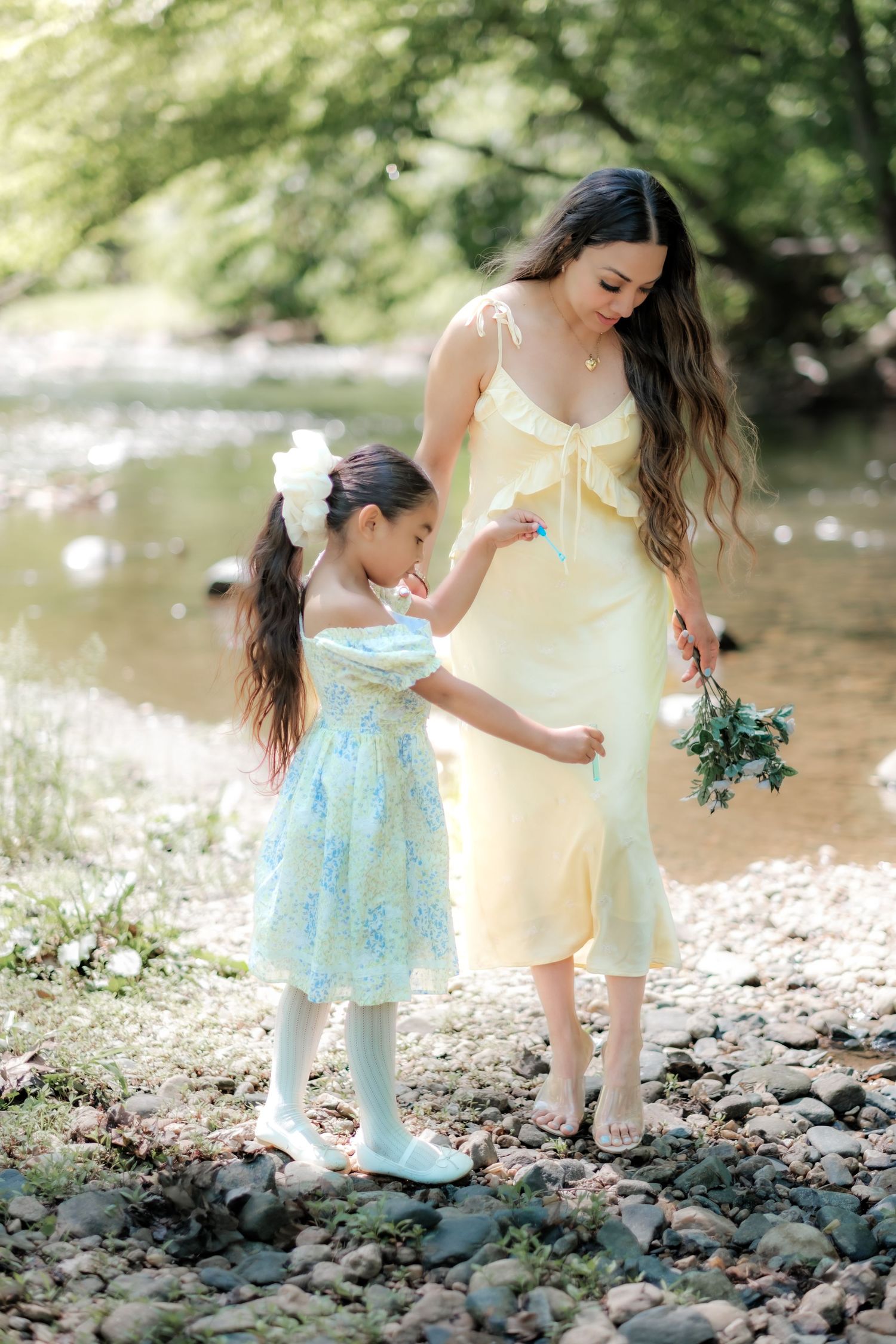 A mother in a yellow dress and her daughter in a blue floral dress walk by a rocky creek.