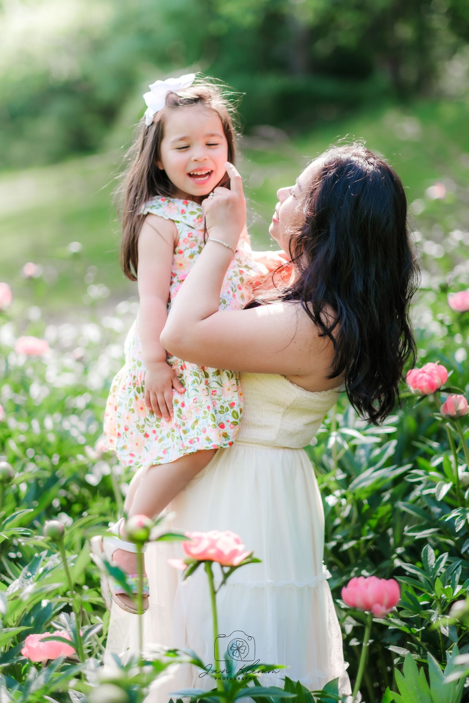 A mother holds her young daughter in a blooming peony garden, both wearing floral dresses on a sunny day.