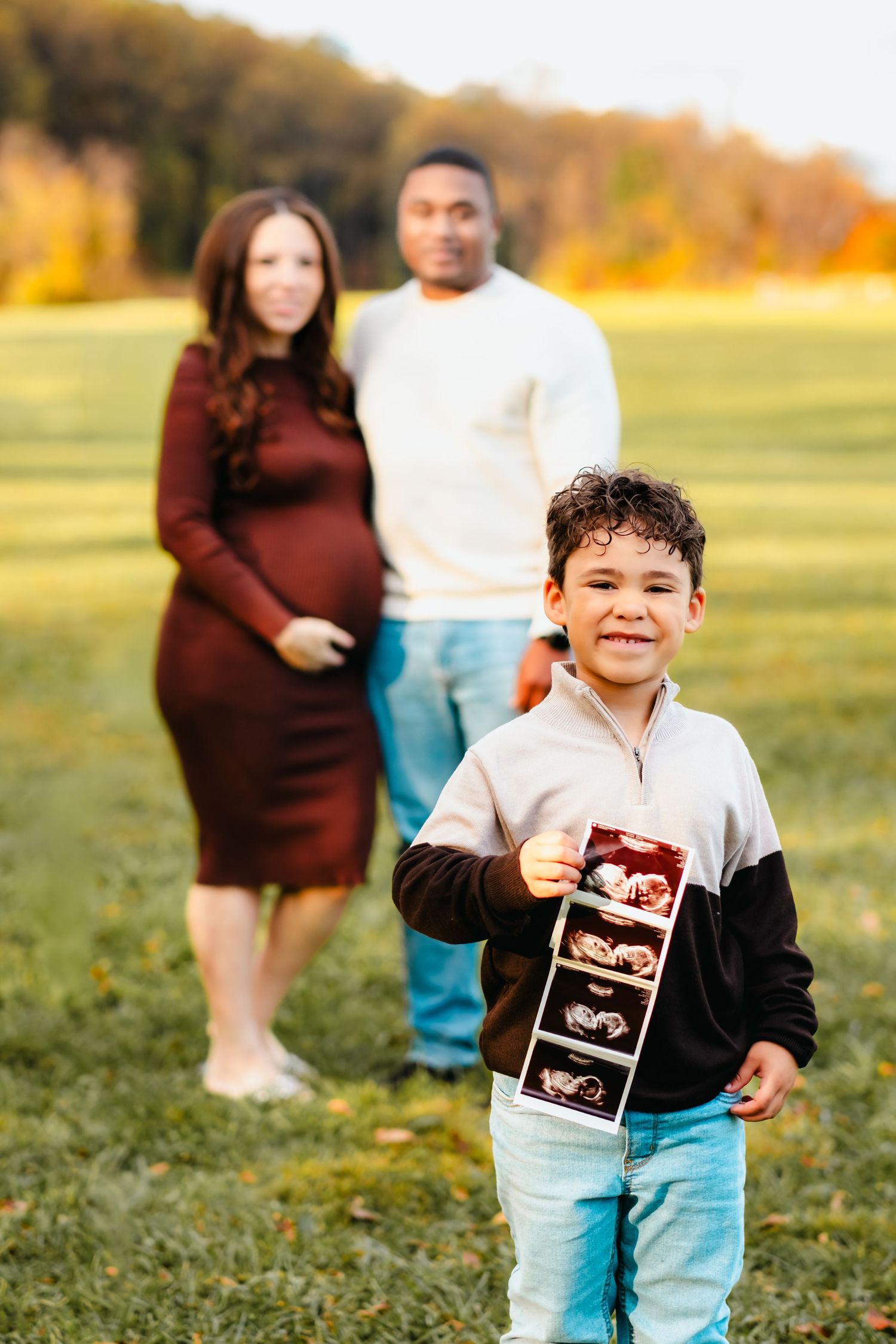 A young boy holds ultrasound photos while his pregnant mother and father stand behind him in a sunny field.