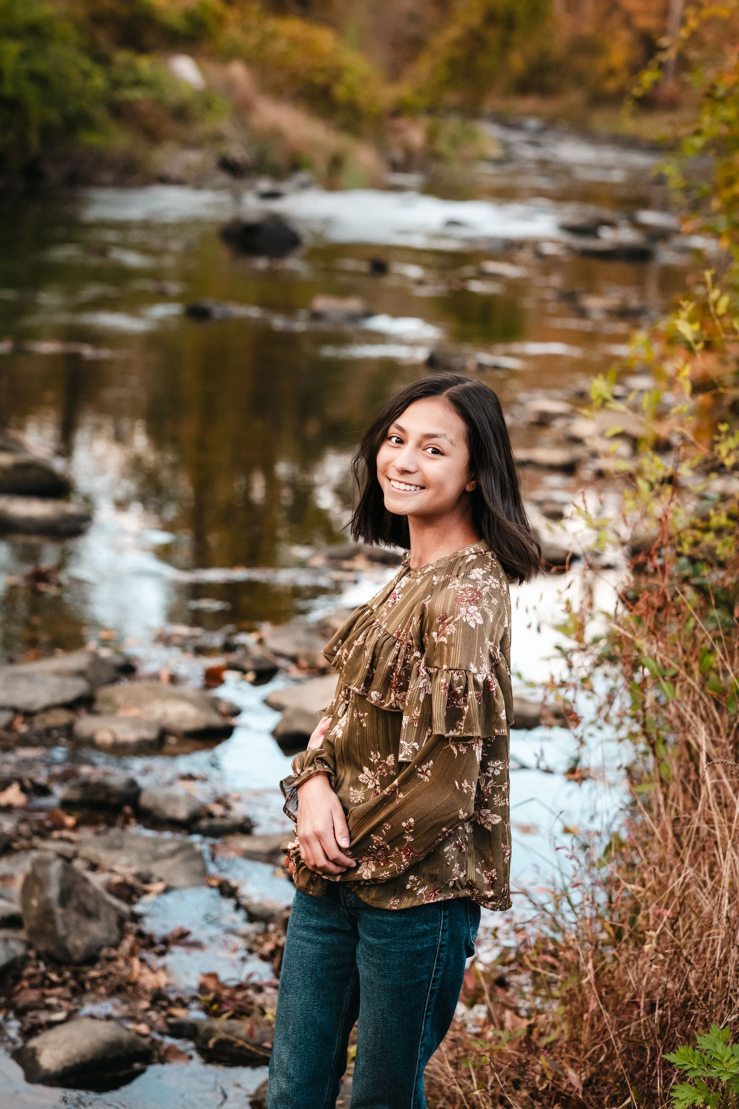 Smiling young woman in floral blouse and jeans poses by a rocky creek surrounded by autumn foliage.
