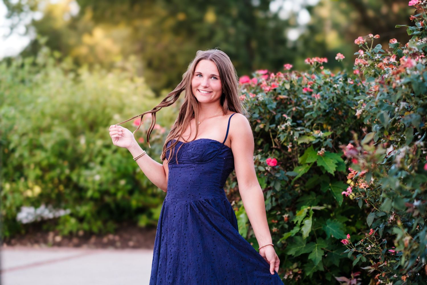 Young woman in navy blue dress twirling hair near rose bushes in a sunny garden.