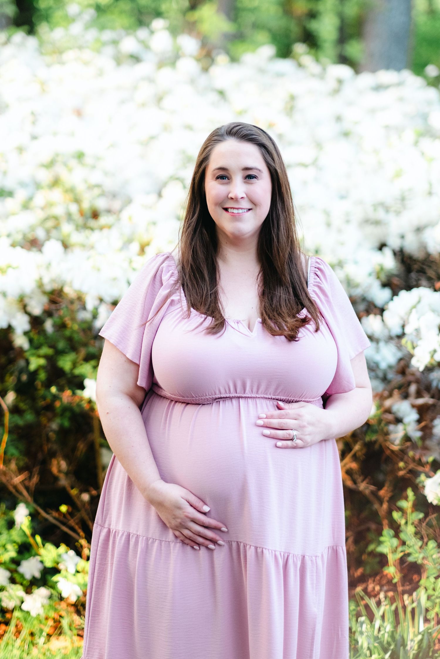 Smiling pregnant woman in pink maternity dress holding baby bump outdoors with white flowering bushes background.
