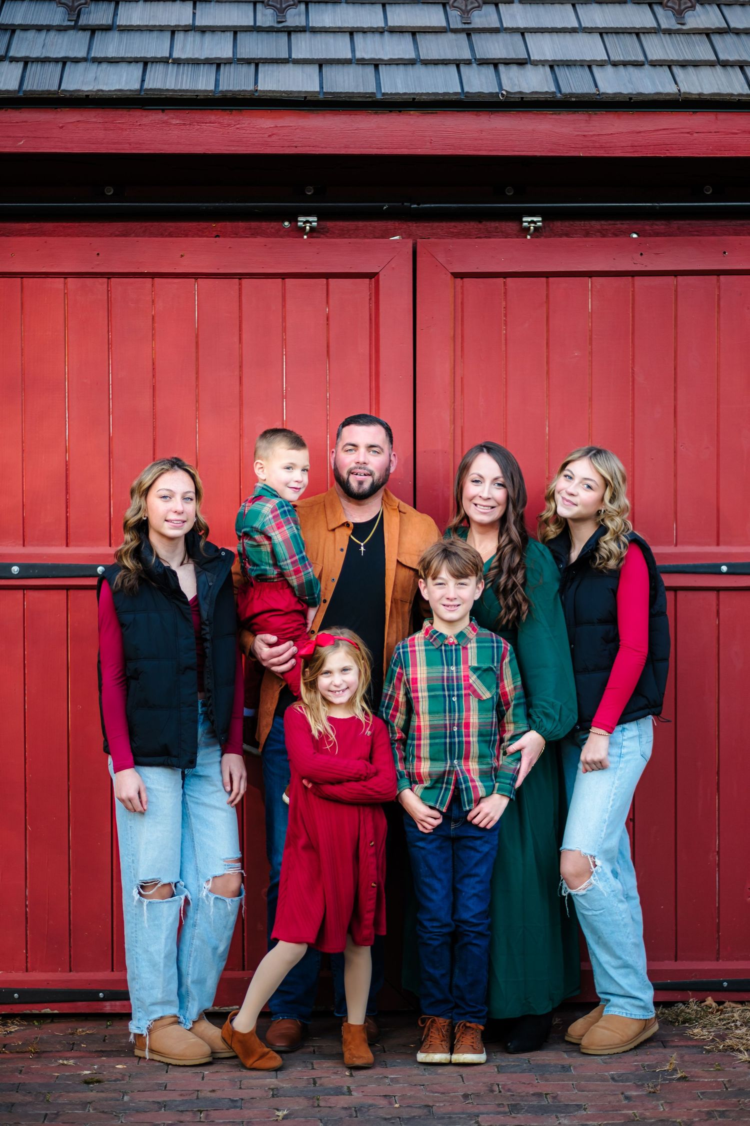 Family of seven posing in festive holiday outfits in front of a rustic red barn door.