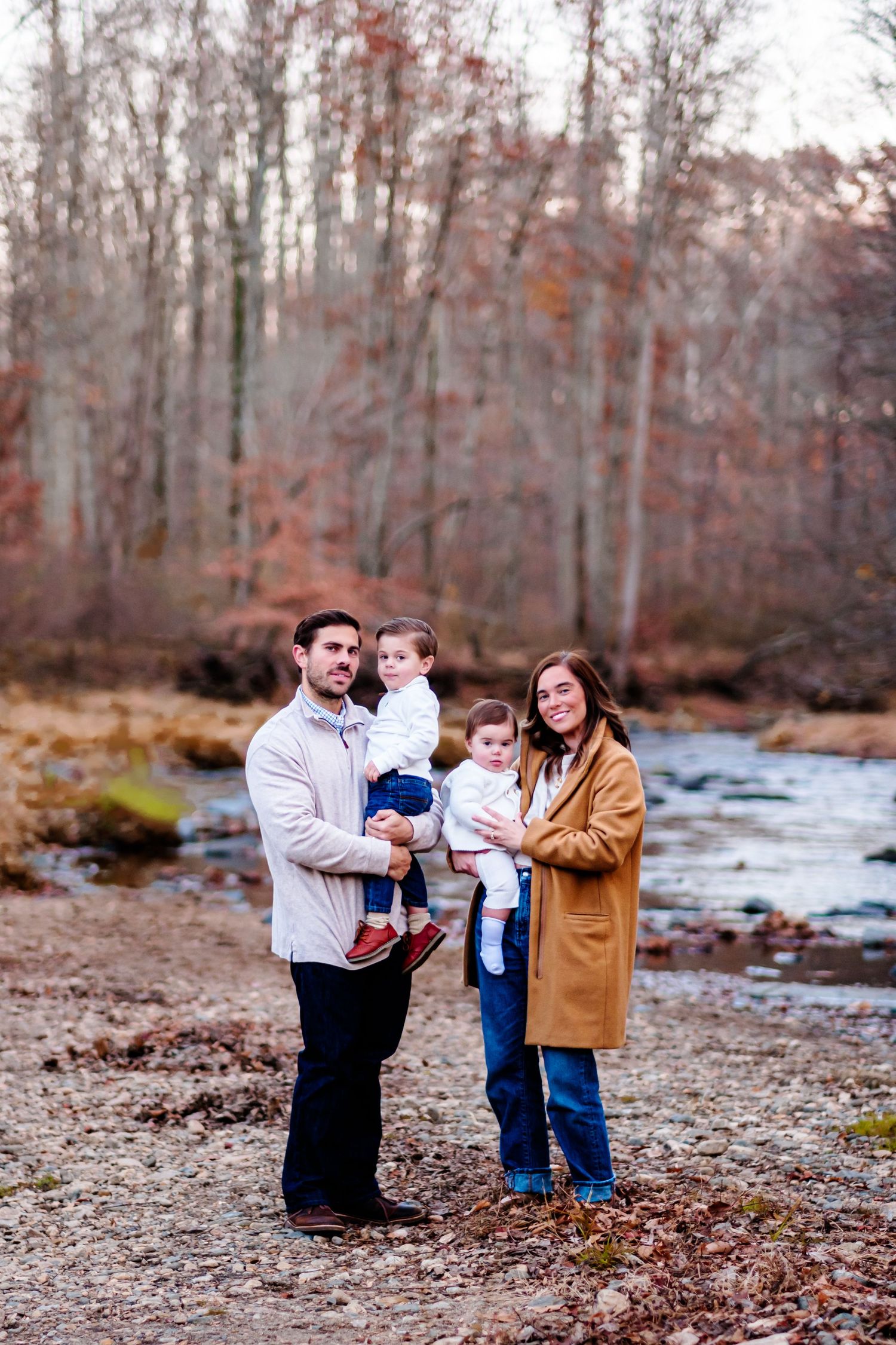 Family of four posing by a creek in autumn woods, parents holding two young children, wearing casual fall outfits.