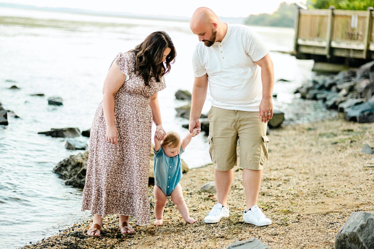 Family of three enjoying a sunny day at a rocky beach shoreline, parents watching toddler explore the water's edge.